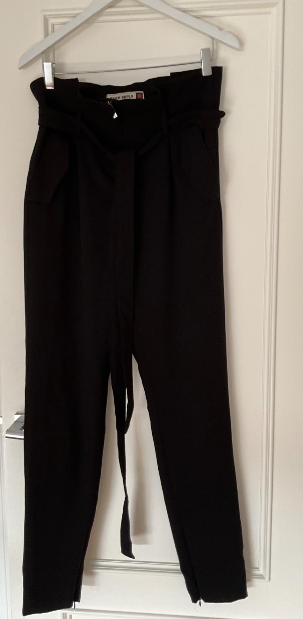 Black trousers hanging on a white hanger above a white door. Label visible with text "ZARA SRPLS".