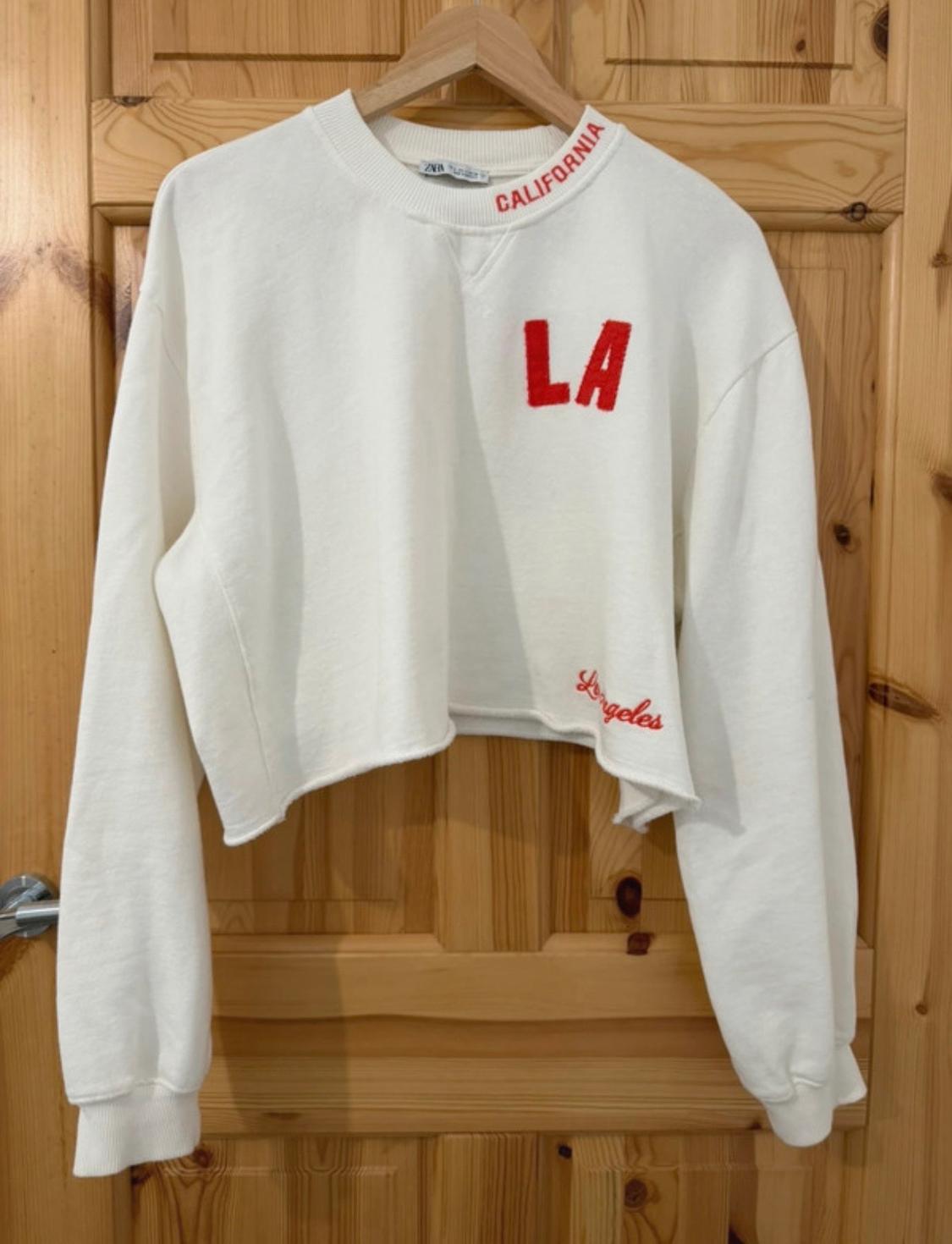 White cropped sweatshirt hanging on a wooden door. Red details: "CALIFORNIA" on the neck, "LA" on the chest and "Los Angeles" on the hem.