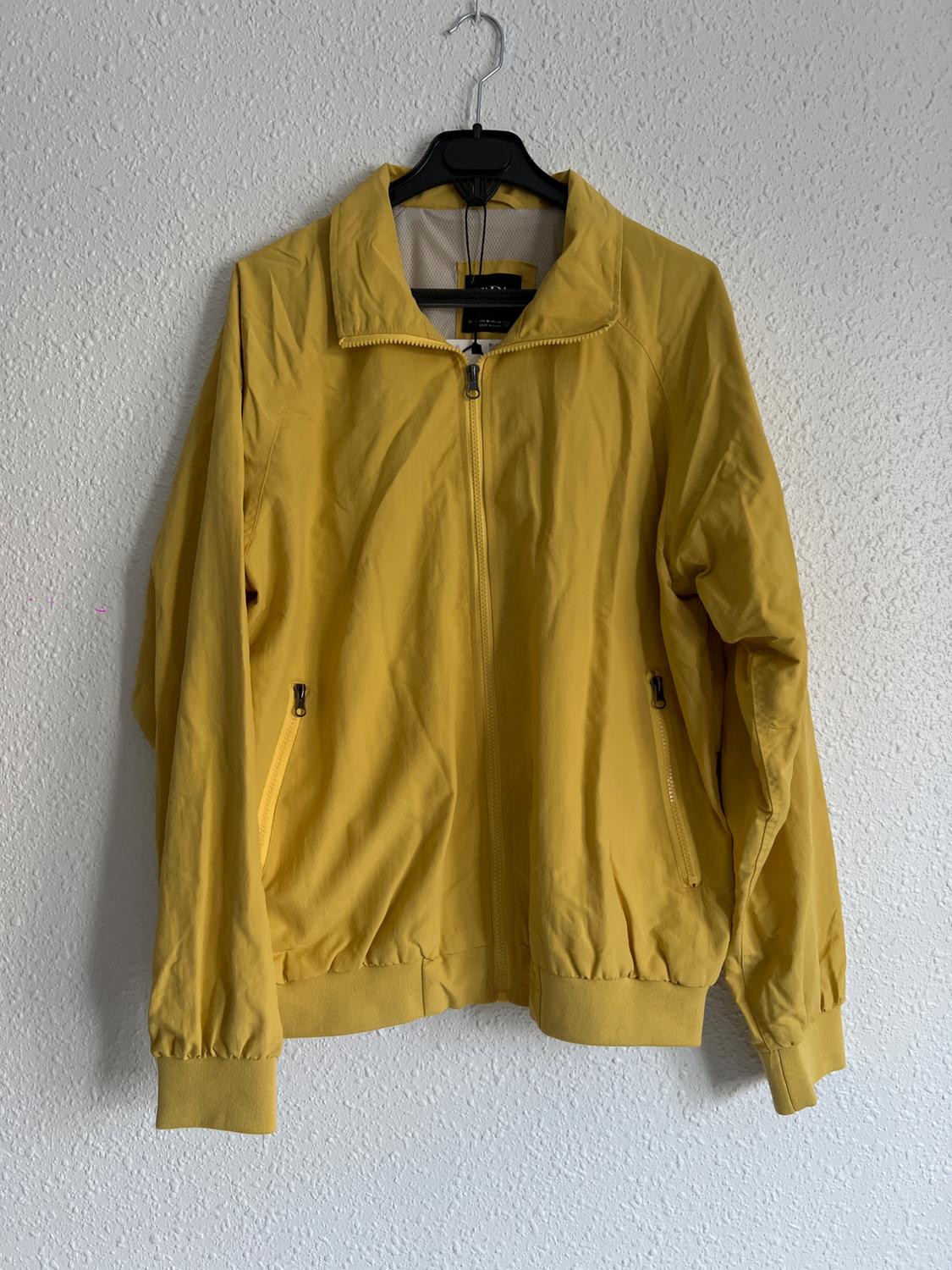 Yellow jacket hanging on a black hanger against a white wall. It has a front zip and side pockets with closures.