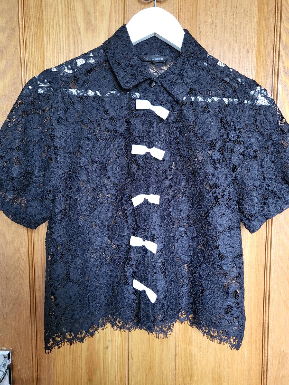 Black lace blouse with short sleeves and white ties on the front, hanging on a hanger against a wooden background.