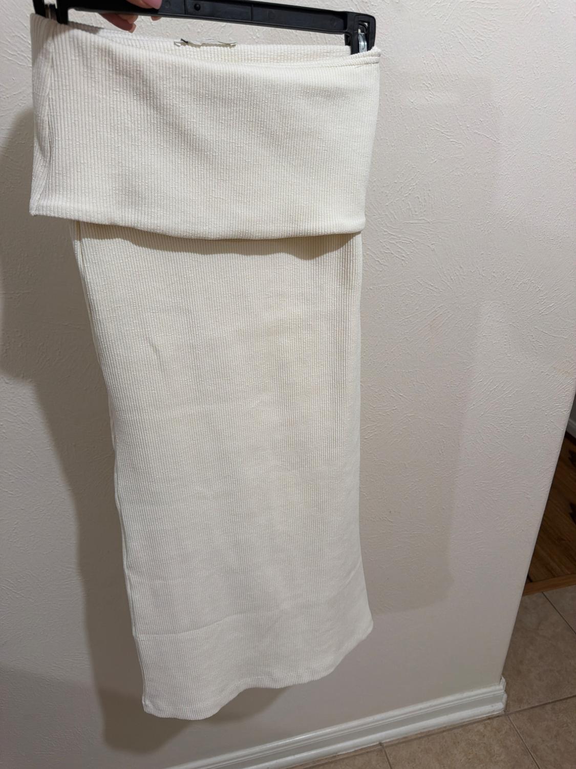 White ribbed knitted garment with a folded top, hanging on a black hanger against a white wall.