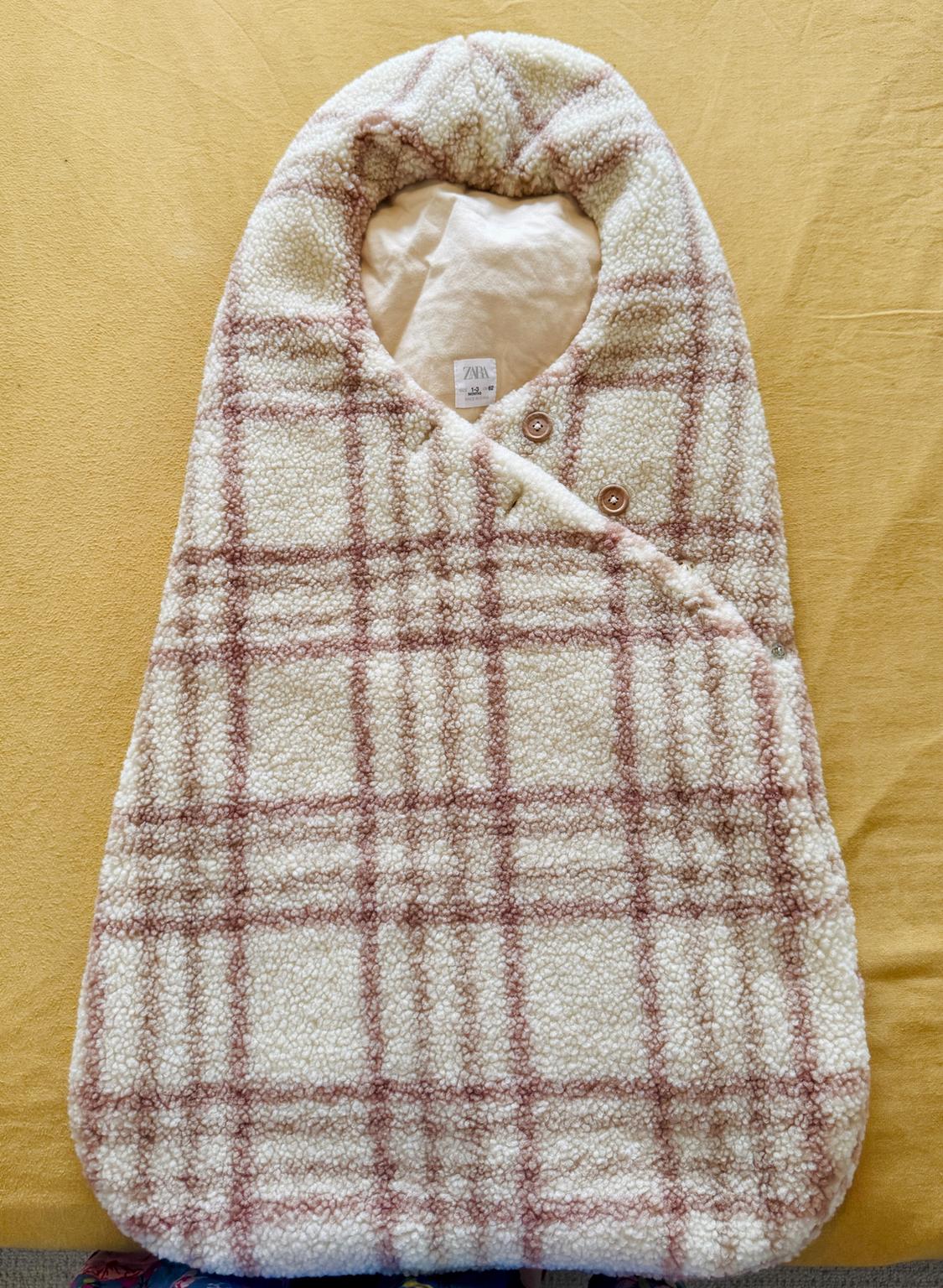 Cream and brown checked baby sack with a plush texture. Crossover design with buttons and a ZARA label inside, on a mustard background.