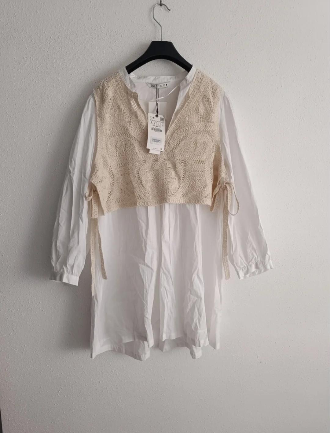 White dress with a beige lace blouse over the top, hanging on a black hanger against a white wall. Tags visible on the collar.