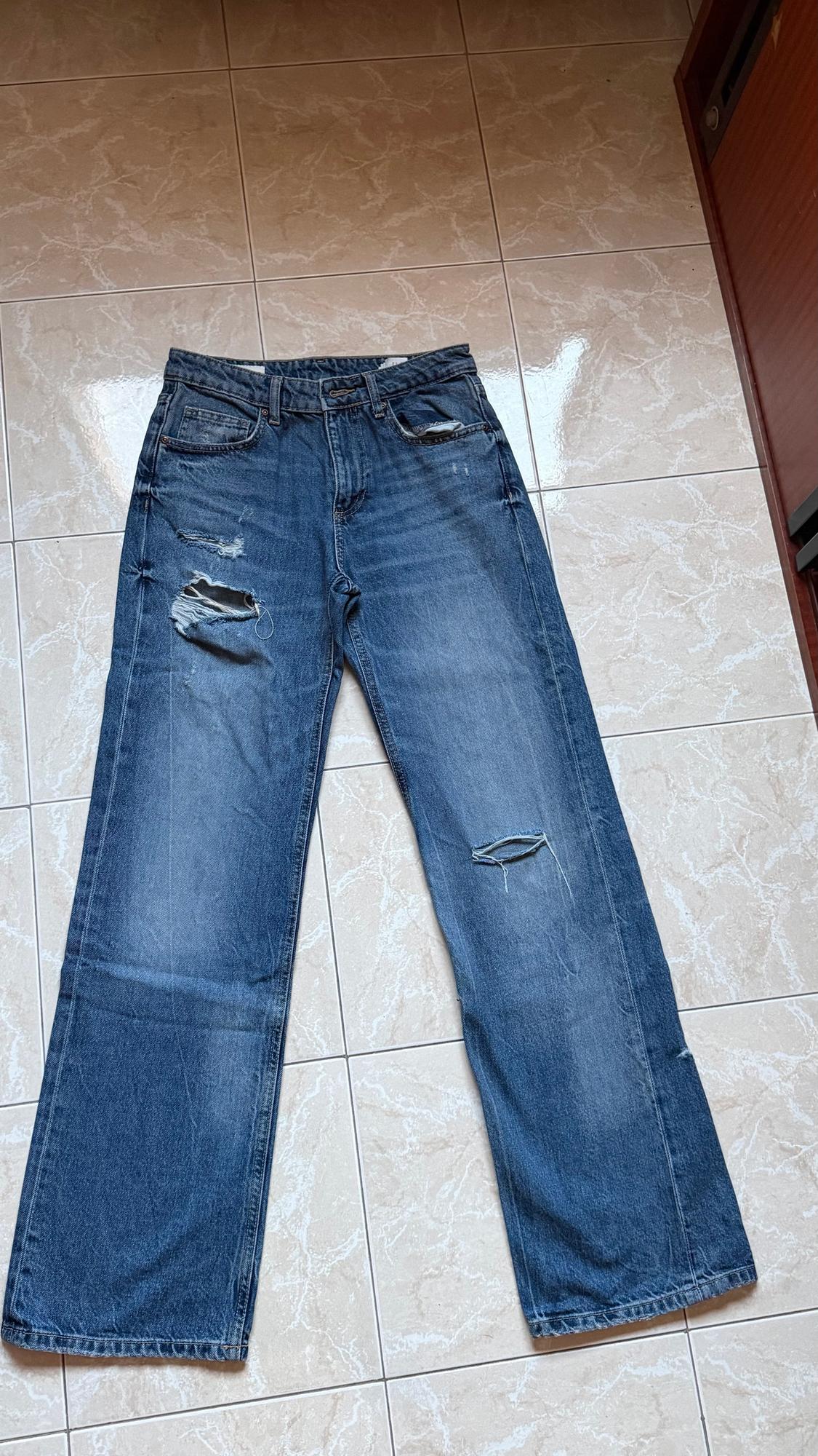 Worn blue jeans with rips on the legs, placed on a light tiled floor.