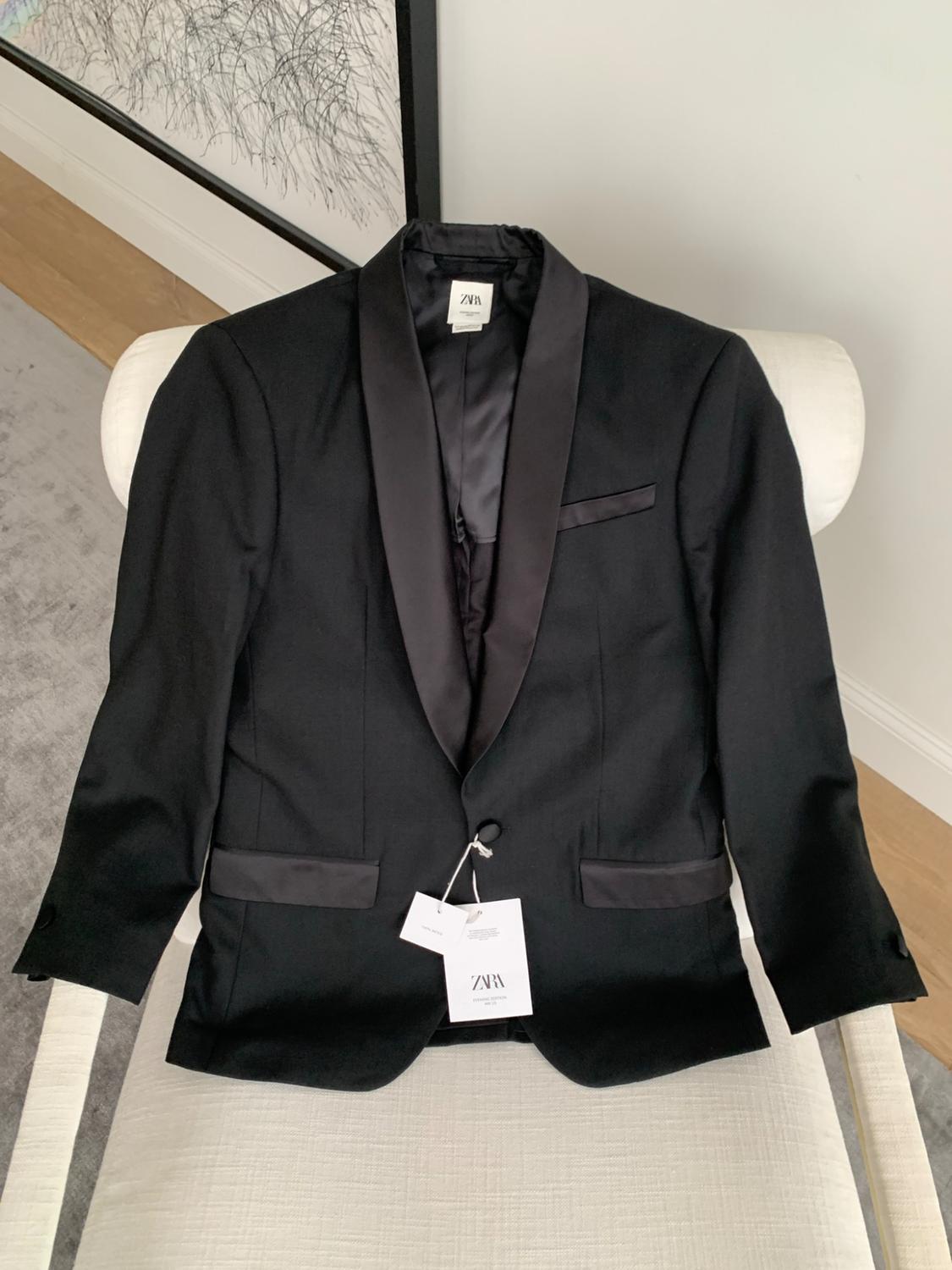 Black jacket with satin lapels on a white chair. Includes labels bearing the text ZARA. Background with a grey rug and abstract painting.