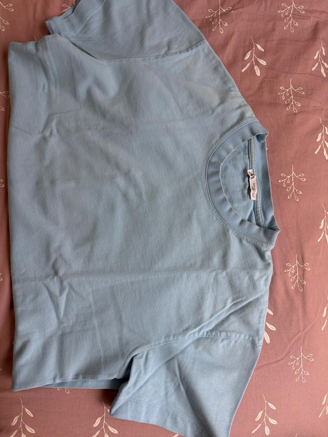 Light blue short-sleeved t-shirt, slightly wrinkled, resting on a pink sheet with a small white leaf pattern.