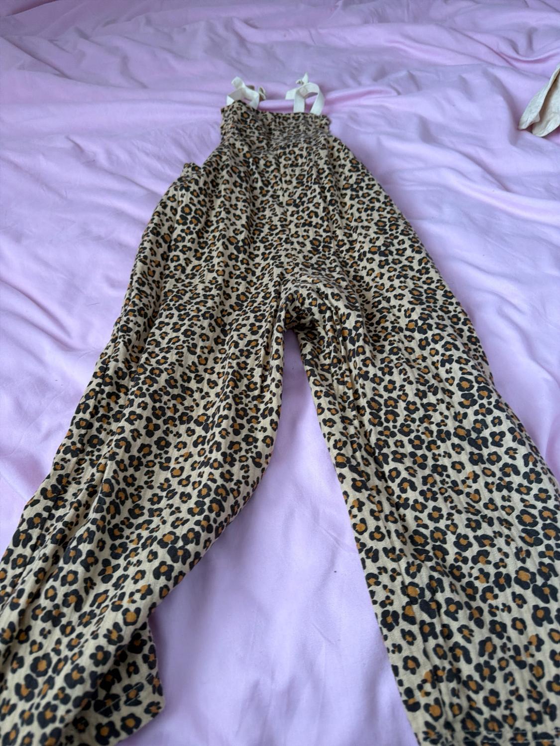 Leopard-print jumpsuit with tied white straps, laid out on a crumpled light pink sheet.