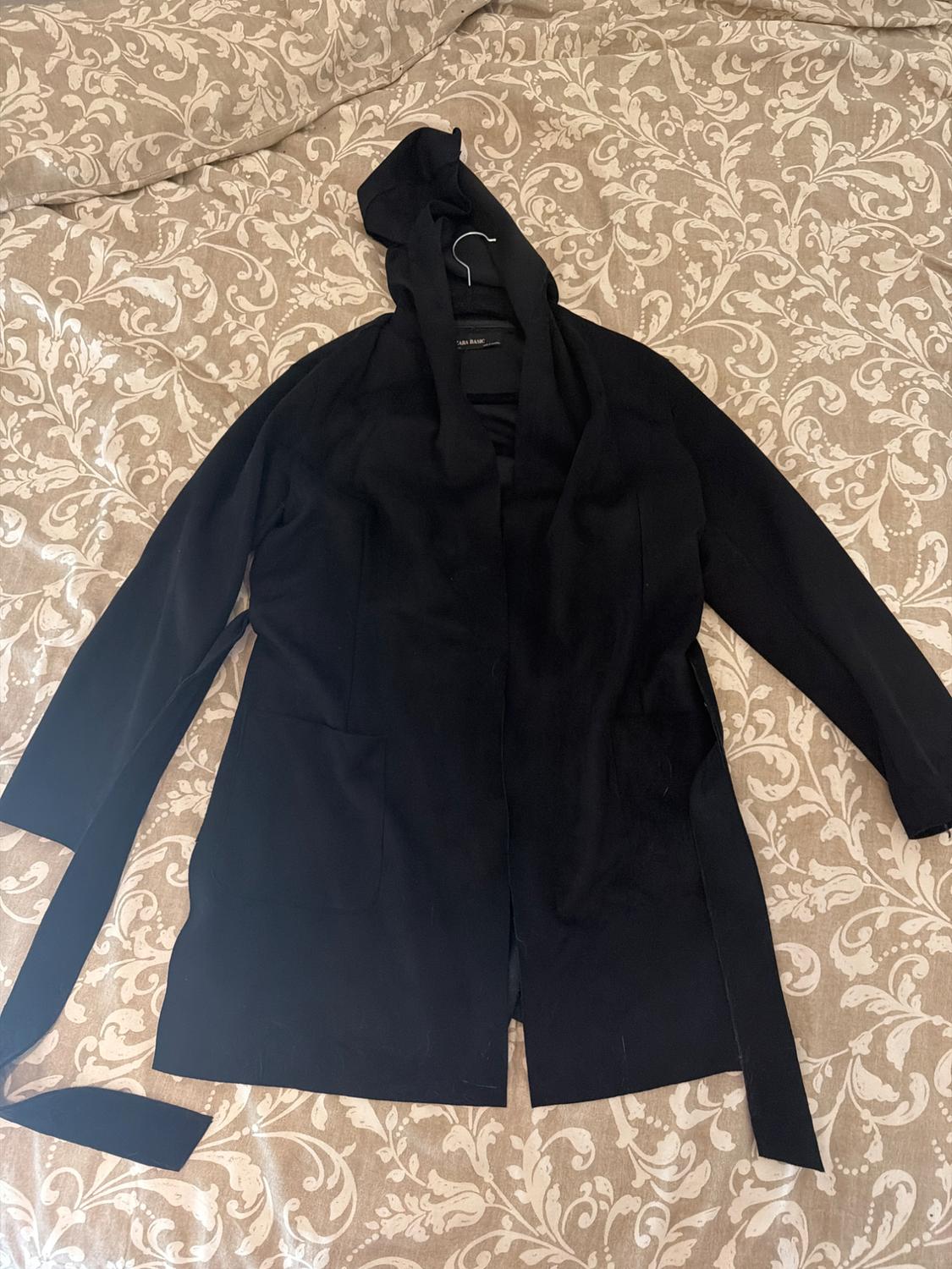 Black hooded jacket with pockets and a belt against a beige floral background. Text: ZARA BASIC.