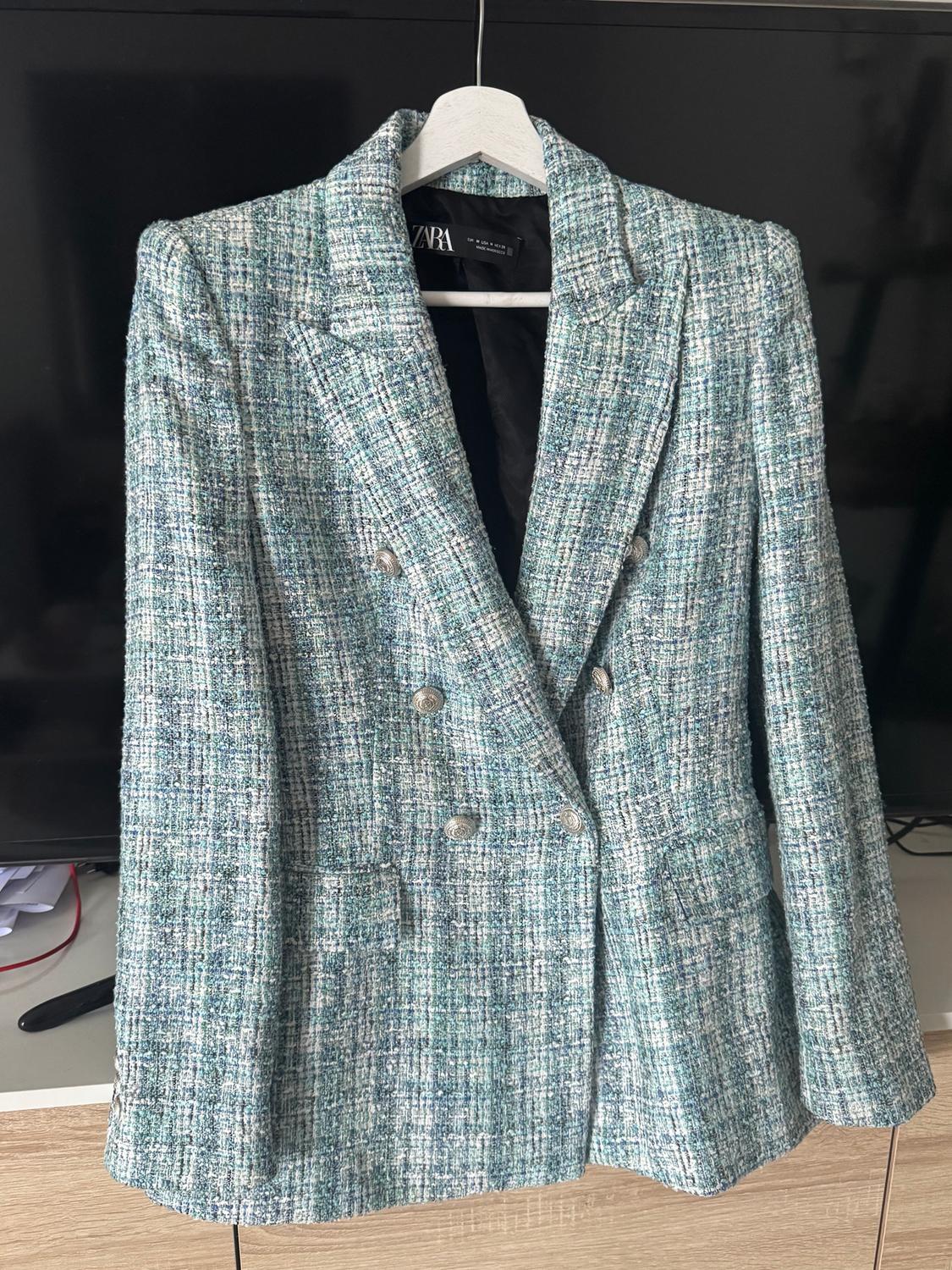 Textured fabric jacket in shades of green and blue, with metal buttons, hanging on a hanger against a dark background.