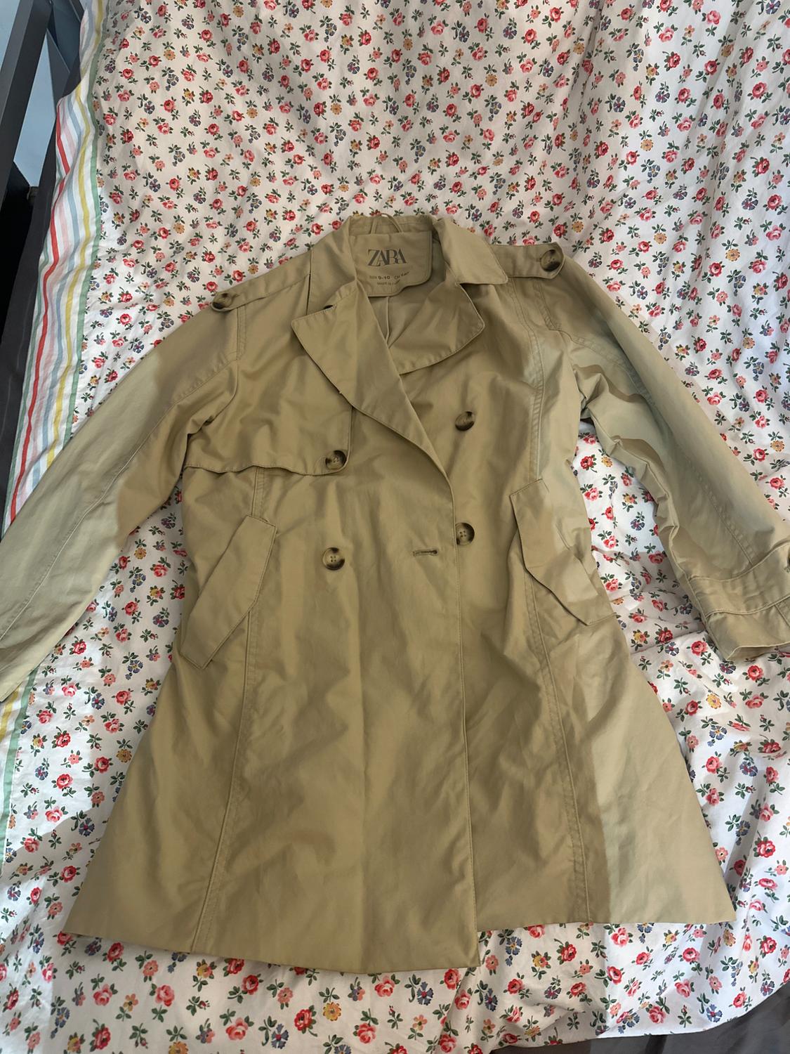 Beige button-up trench coat laid out on a floral print sheet. The label inside reads ZARA.