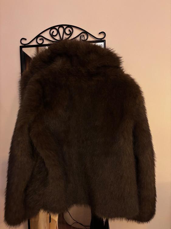 Brown fur coat hanging on a black metal coat rack with a decorative design at the top, against a beige wall.