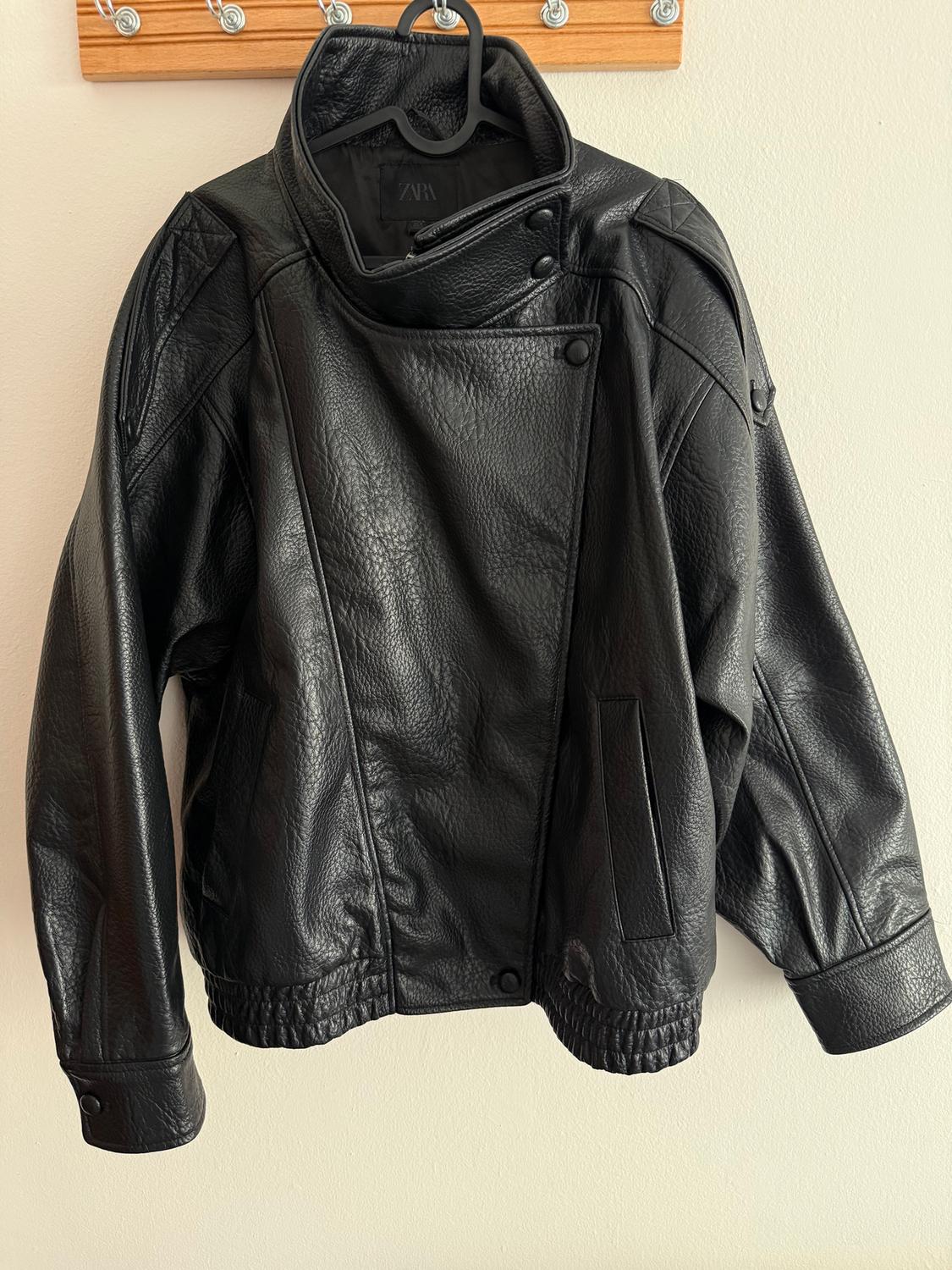 Black leather jacket hanging on a black hanger against a light background. Design with a high collar and visible buttons.