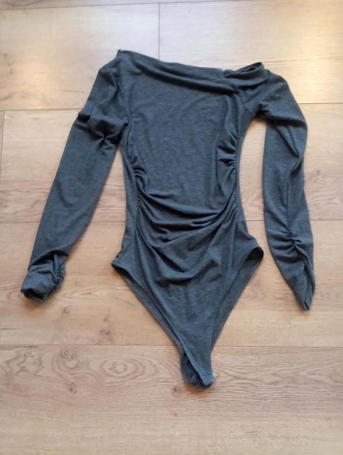 Dark grey, long-sleeved bodysuit with an asymmetrical neckline and ruching on the sides and sleeves, displayed on a light wooden floor.