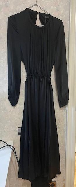 Long-sleeved black dress hanging on a hanger against a light-coloured wall. Elegant design with an elasticated waist and round neck.