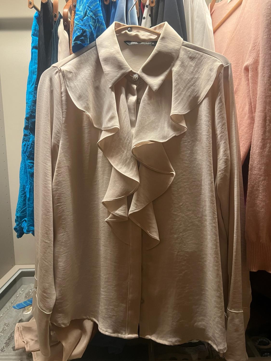 Beige long-sleeved blouse with front frills, hanging in a wardrobe alongside other garments. ZARA text visible on the label.