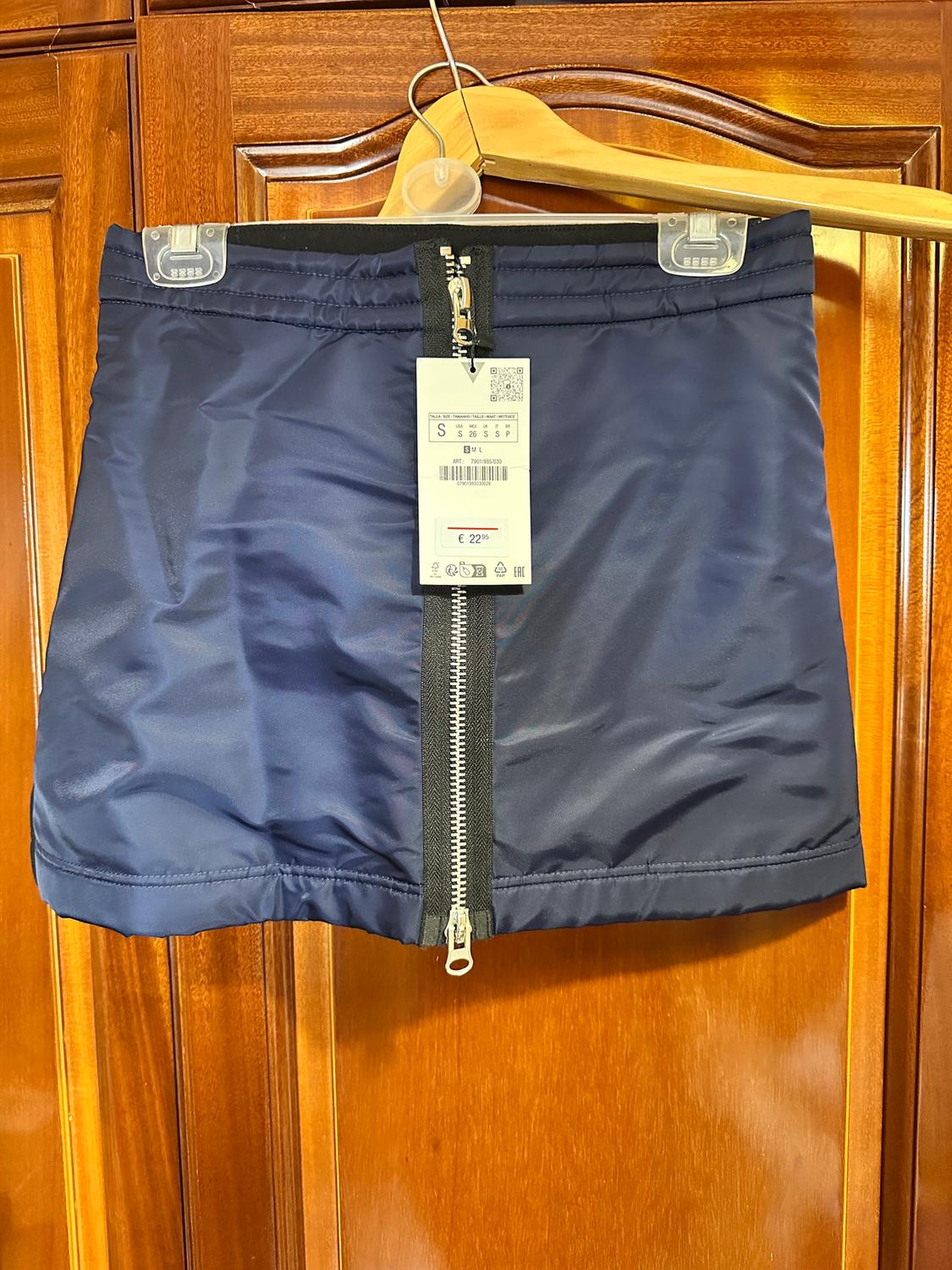 Blue skirt with front zip hanging on a wooden hanger. Visible tag with price of €22.99 and size S. Wooden background.