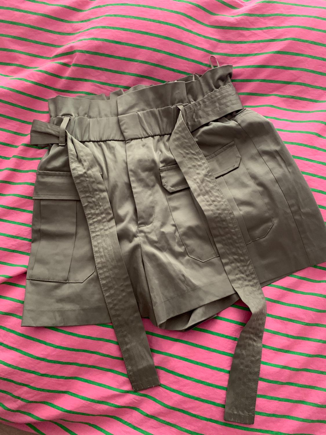 Khaki high-waisted ruched shorts with a belt and pockets, on a pink fabric background with green stripes.