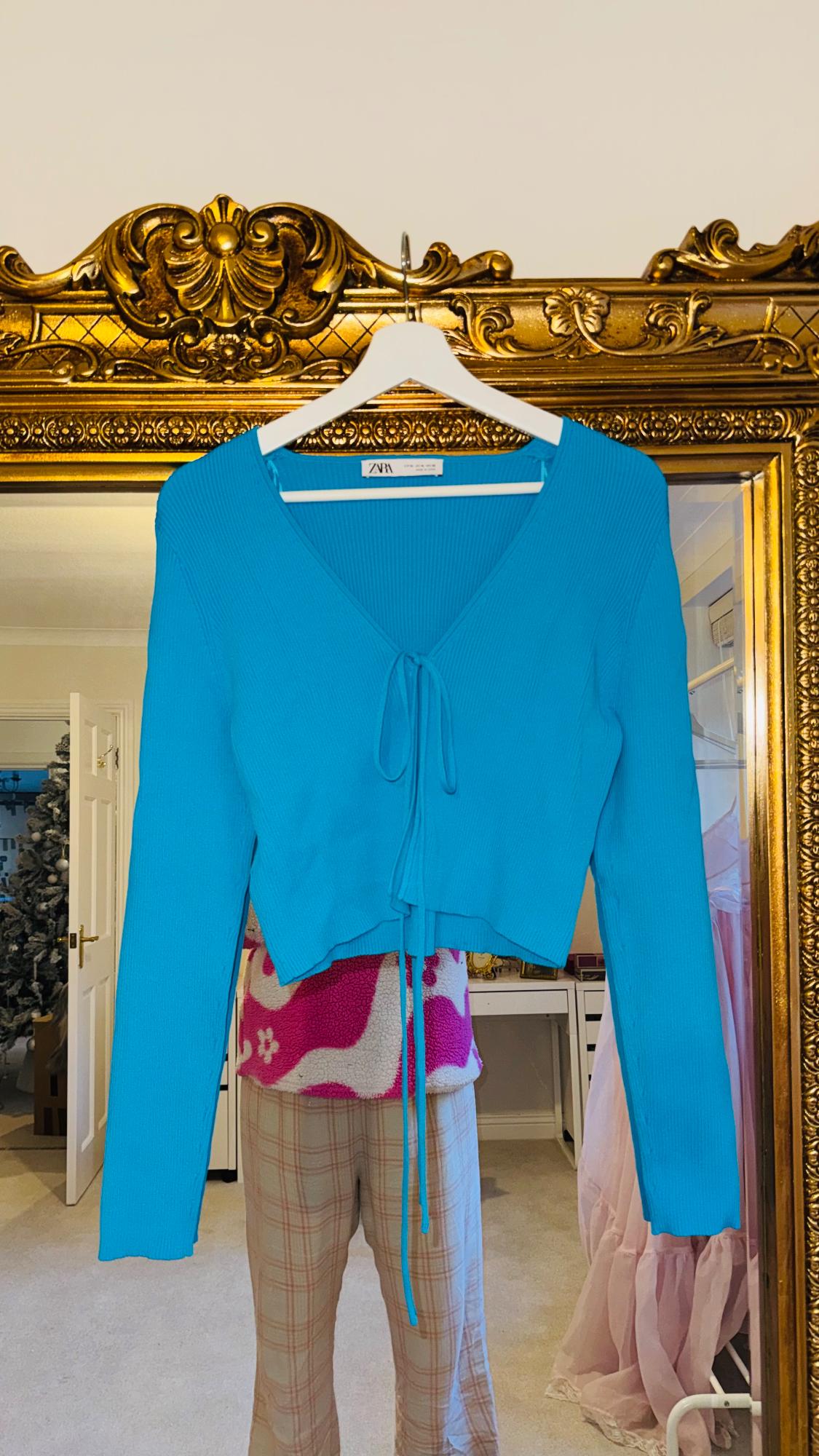 Blue cardigan with a bow hanging on a white hanger in front of a gold mirror. Background with Christmas decorations and clothes.