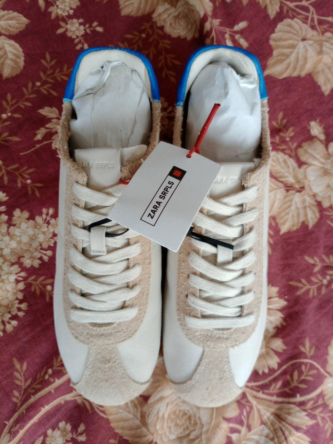 White and beige trainers with blue detail on a red floral background. Label featuring the text ZARA SRPLS.