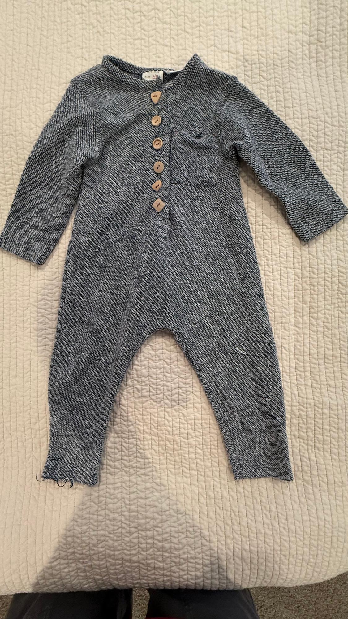 Grey baby romper with wooden buttons and a chest pocket, on a quilted beige textured blanket.