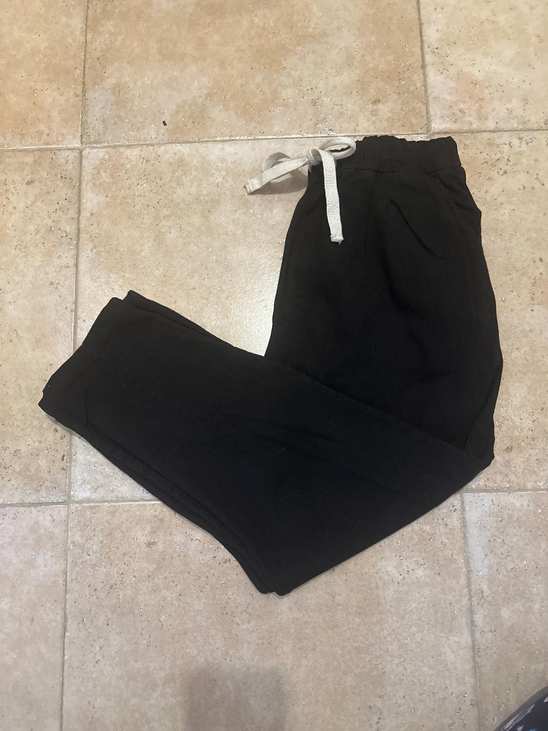 Black tracksuit bottoms with an elasticated waist and white drawstring, placed on a beige tiled floor.