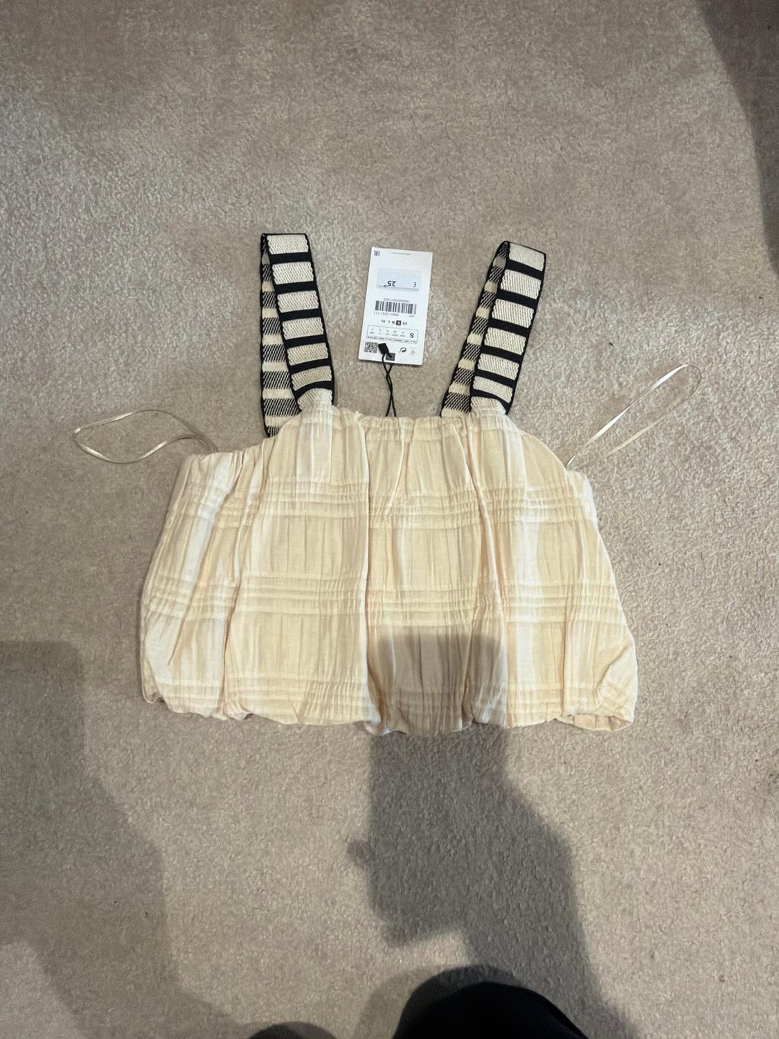 Beige top with black and white striped straps on a grey rug. Hanging tag visible in the centre.