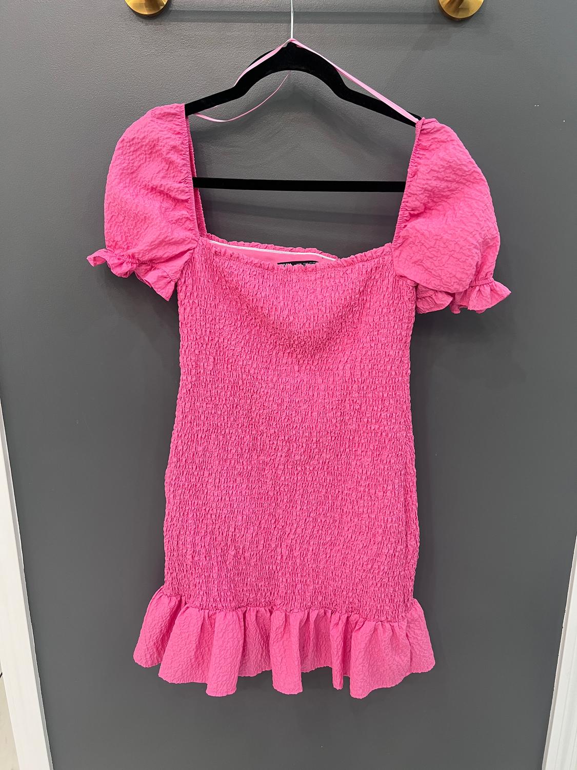 Pink dress with crinkled texture, puffed sleeves, and ruffles on the hem, hanging on a black hanger against a gray background.