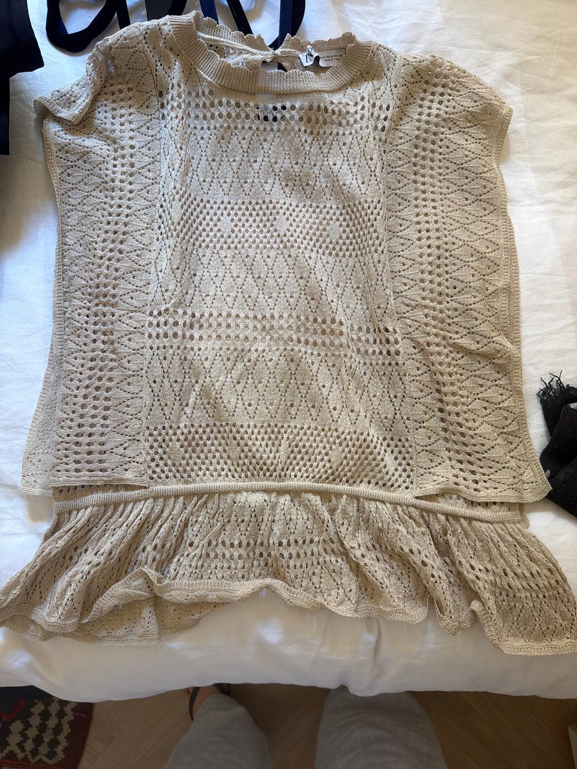 Beige lace blouse with a geometric pattern and ruffles on the hem, on a white bed. Visible tag on the collar.