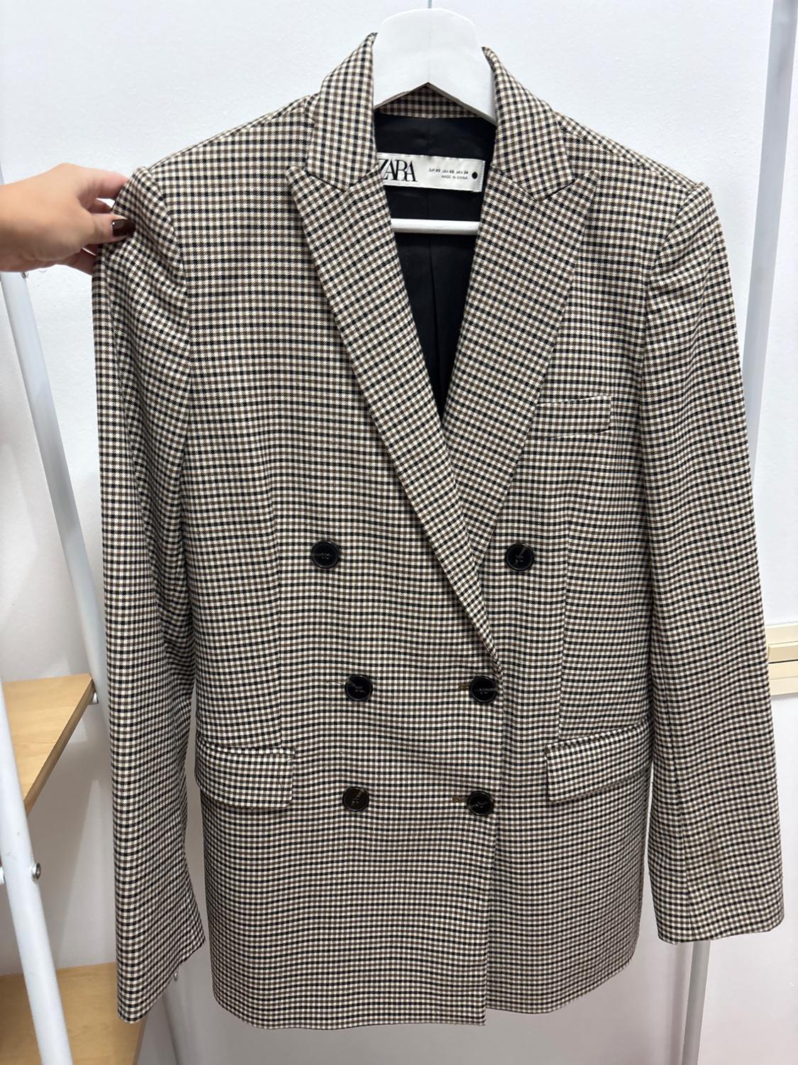 A checked jacket in shades of brown and white is hanging on a white hanger. A hand is holding the left sleeve, and the ZARA label is visible.