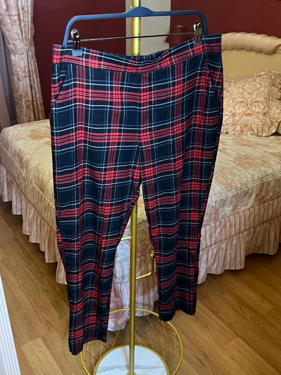 Red and black checked trousers hanging on a gold hanger, against a bedroom background with a bed and curtains.