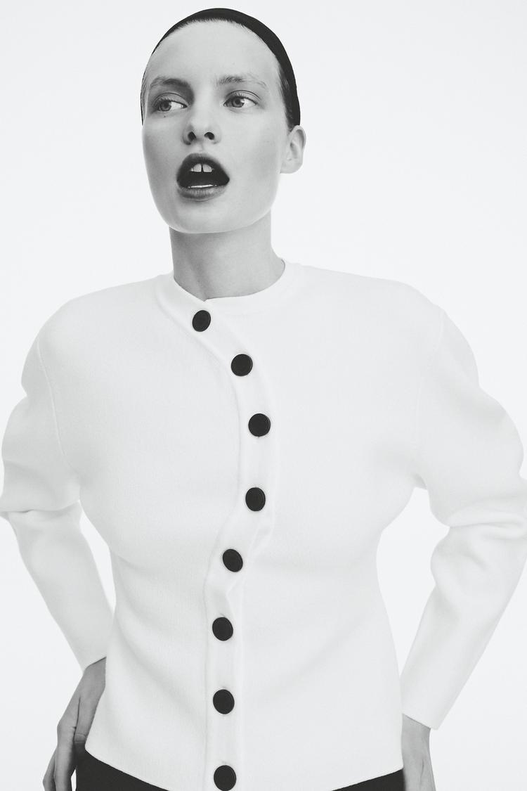 Střední záběr bílé bundy s černými knoflíky v zakřivené linii.