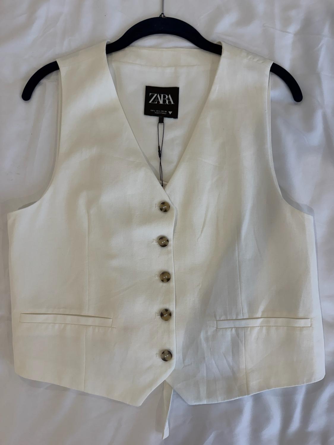 White vest with a V-neck, brown buttons and pockets, on a black hanger against white fabric. Tag with the text ZARA.