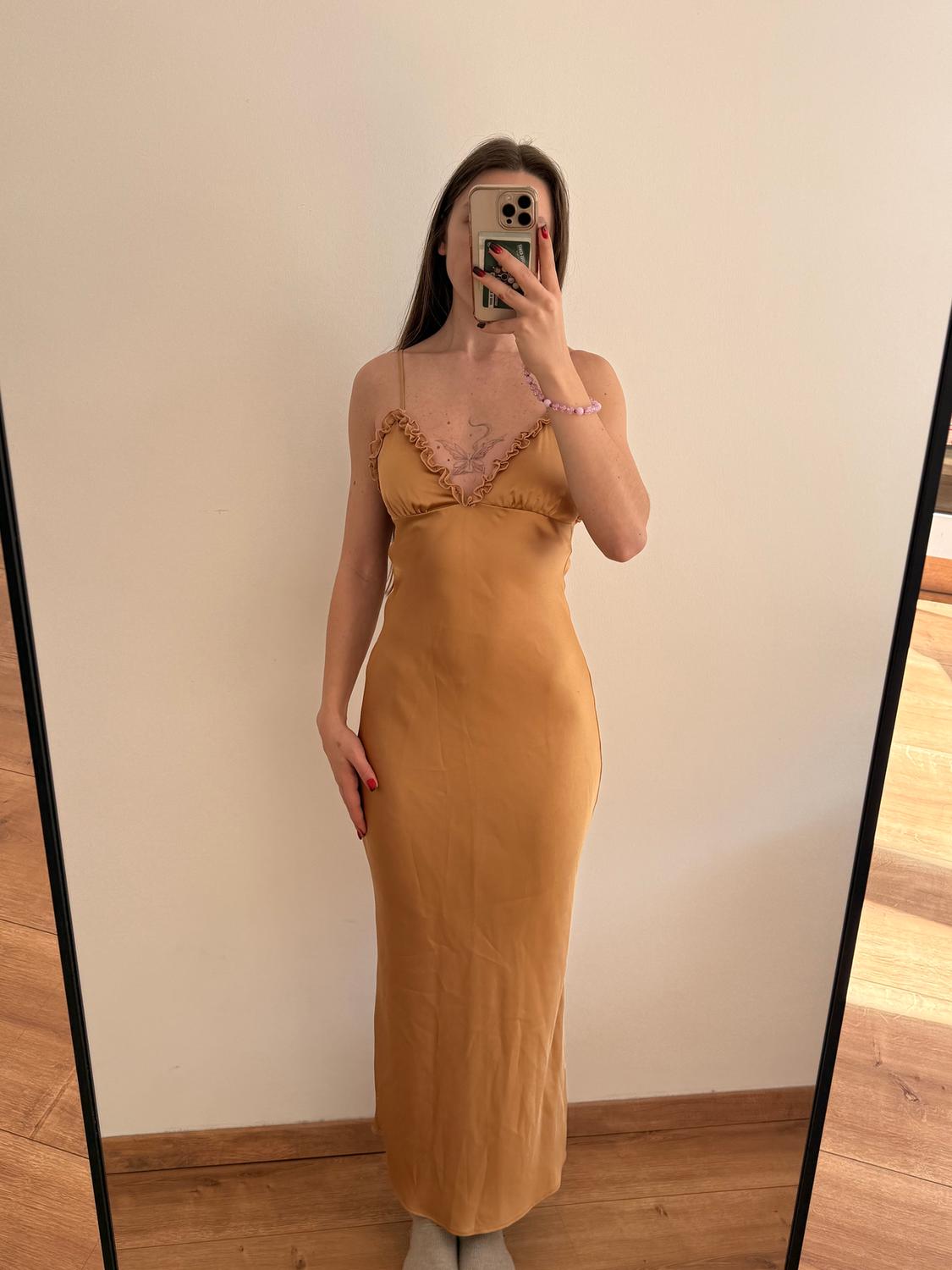 Woman in front of a mirror, wearing a long mustard-coloured dress with thin straps. She's holding a mobile phone, wearing a pink bracelet, and has red nails.