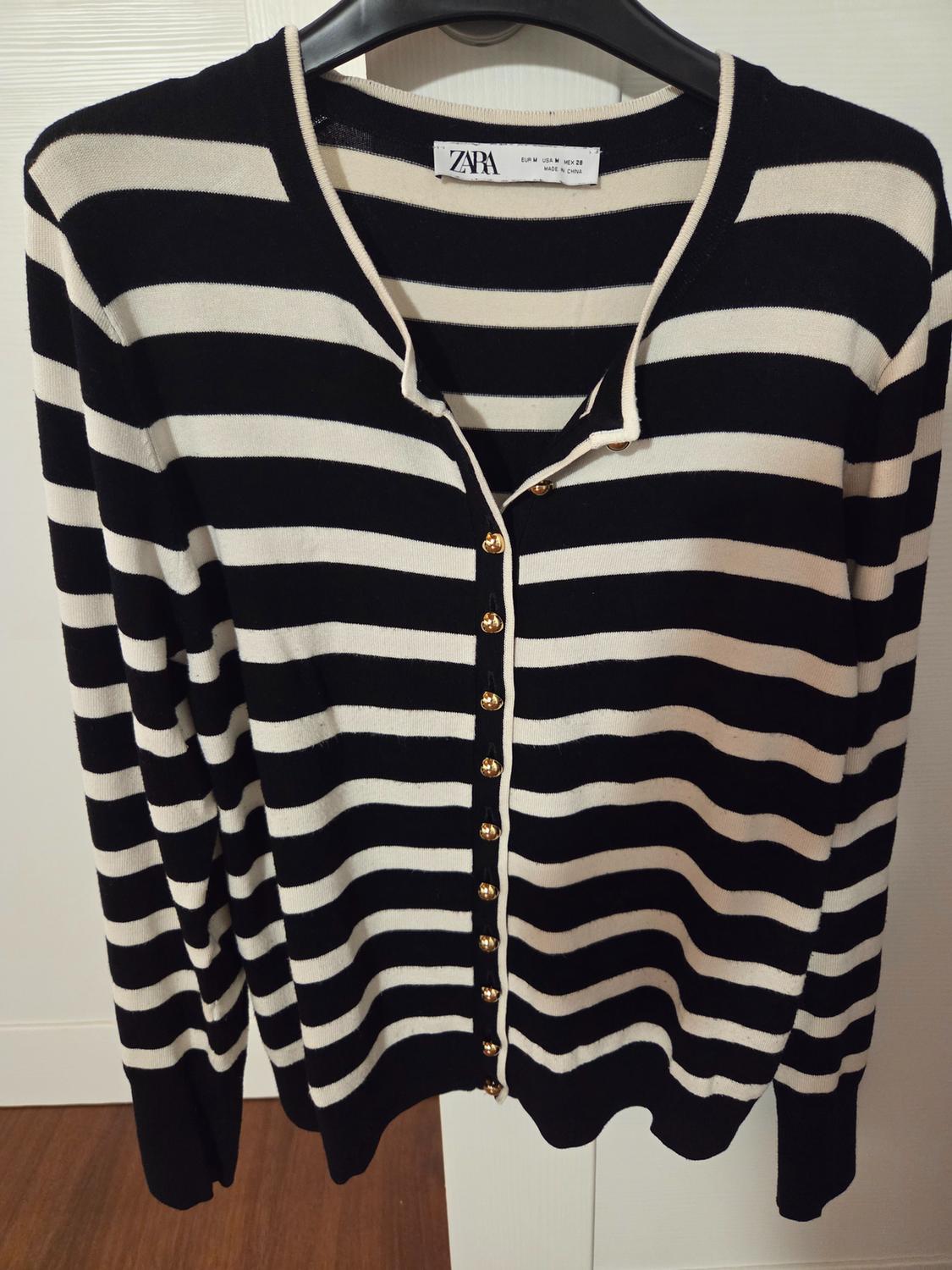 Black and white striped cardigan with gold buttons, hanging on a hanger. Visible label: ZARA, EUR L USA M MX 28, MADE IN CAMBODIA.