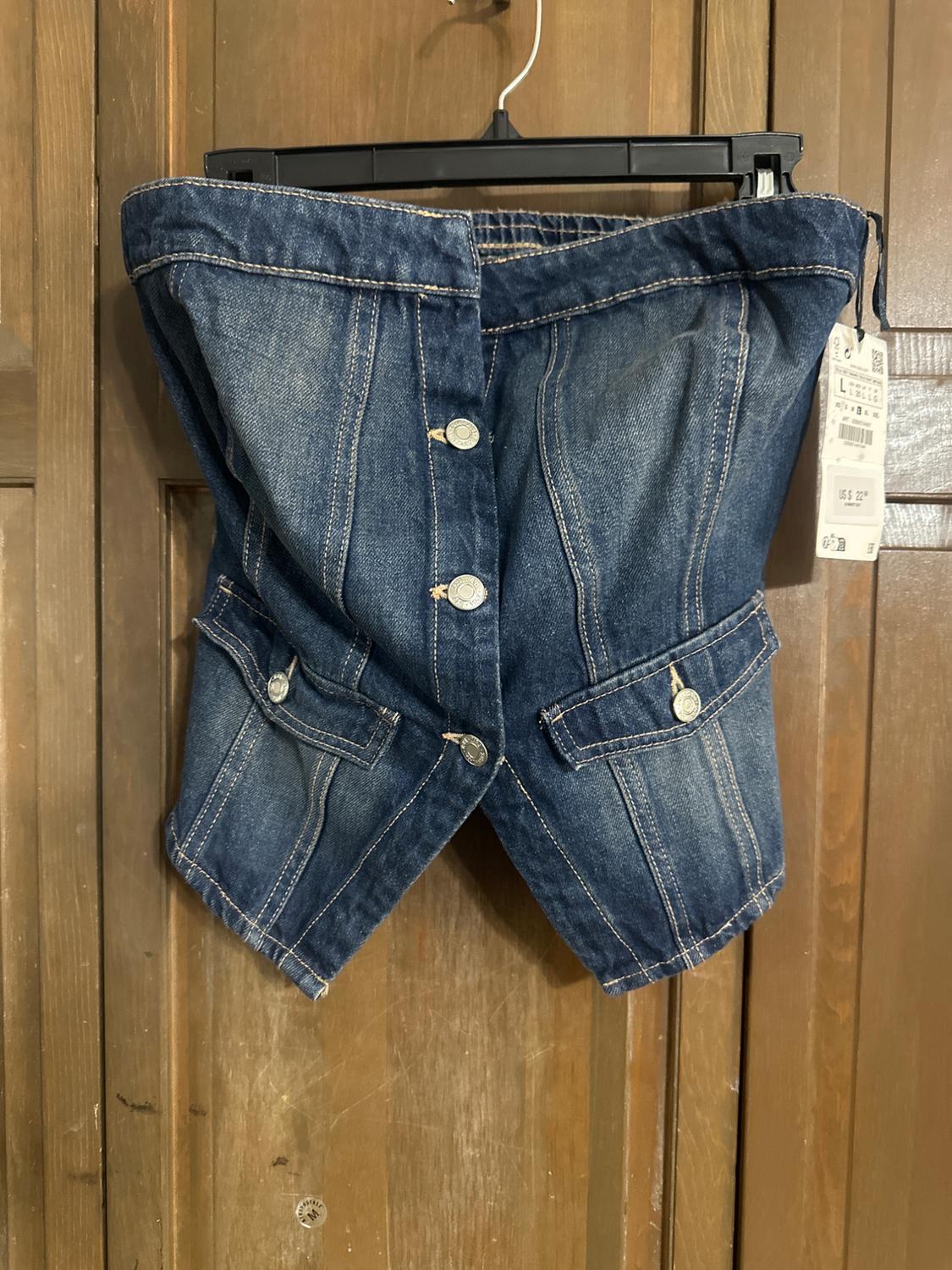 Blue denim top with buttons and pockets, hanging on a black hanger in front of a wooden door. Label visible on the right.