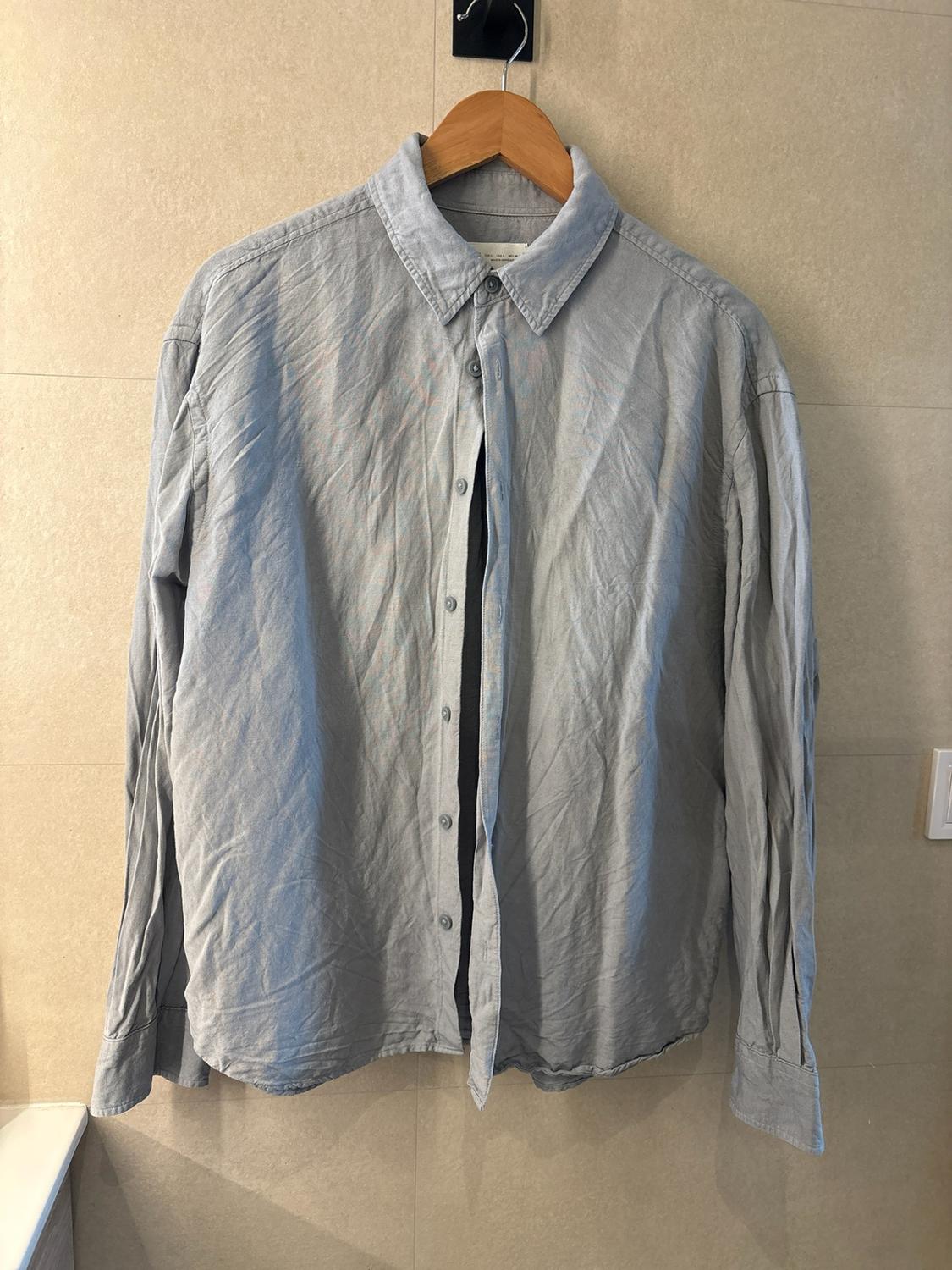 Light grey long-sleeved casual shirt, with a crinkled look, hanging on a wooden hanger against a beige tiled wall.