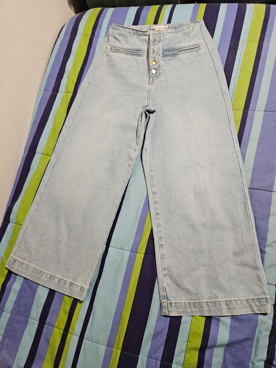 Front view of light blue denim jeans with metal buttons and decorative front pockets.