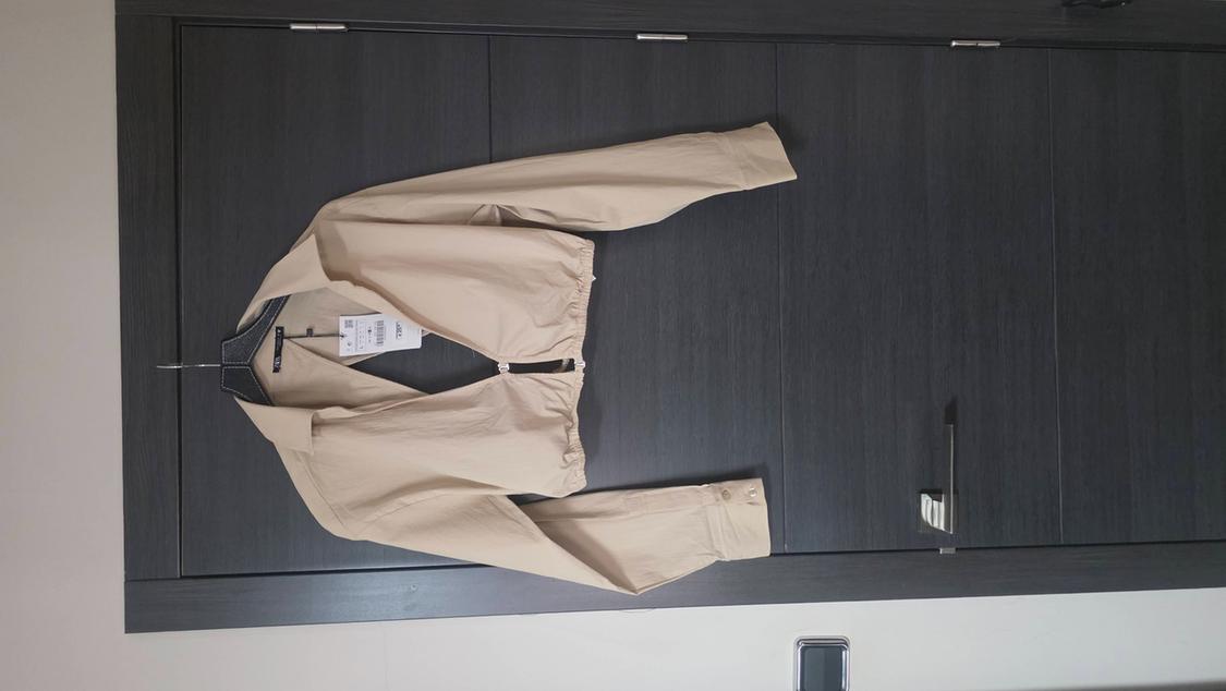 Beige long-sleeved cropped top with a collar and gathered hem, hanging on a hanger on a dark wooden door.