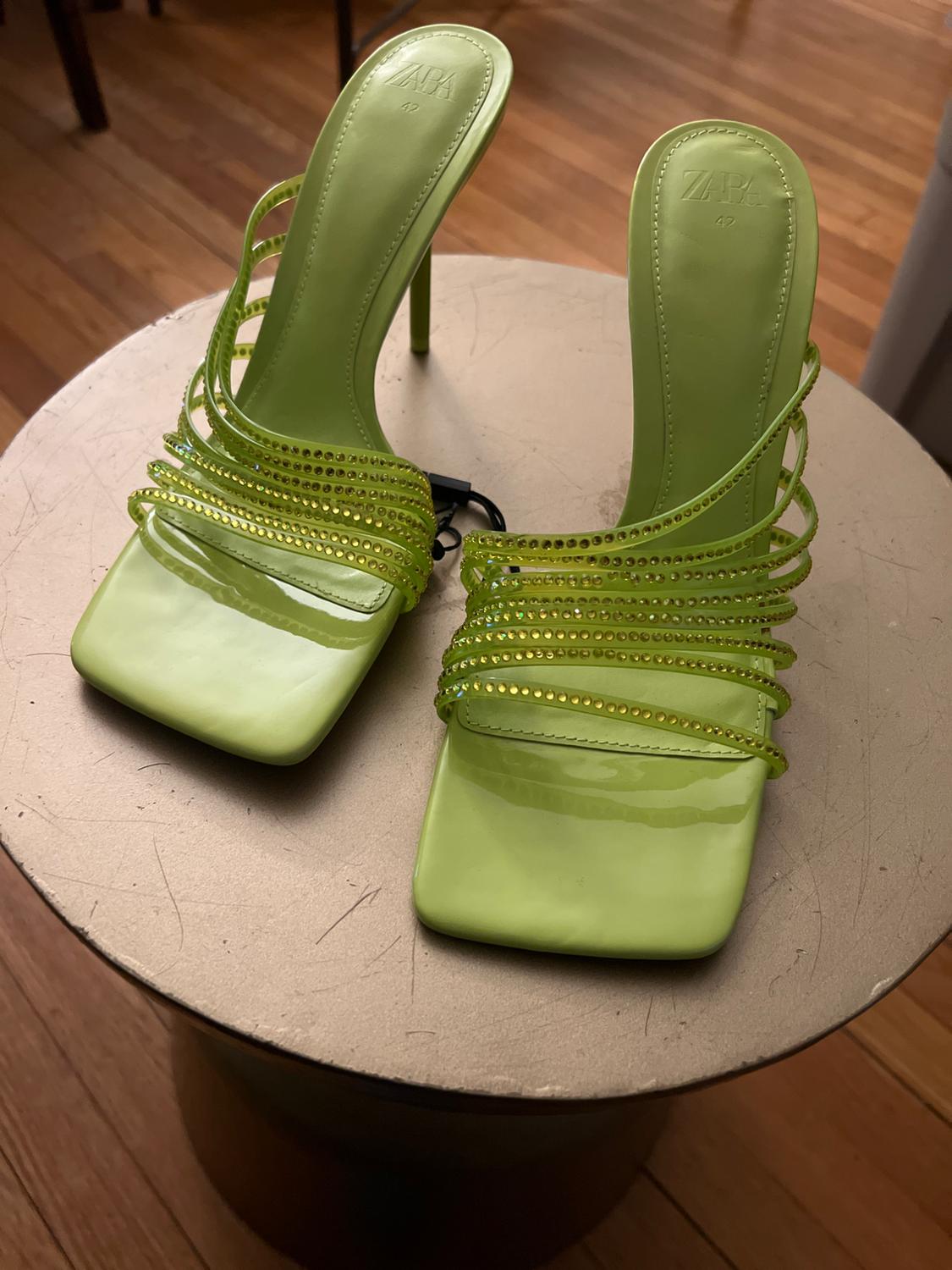 Green high-heeled sandals with sparkly straps on a round table. Visible text: "ZARA 42". Wooden floor in the background.