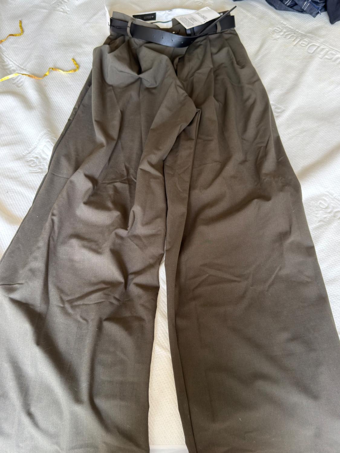 Dark brown wide-leg trousers with a black belt and tags, casually laid out on a white bed.