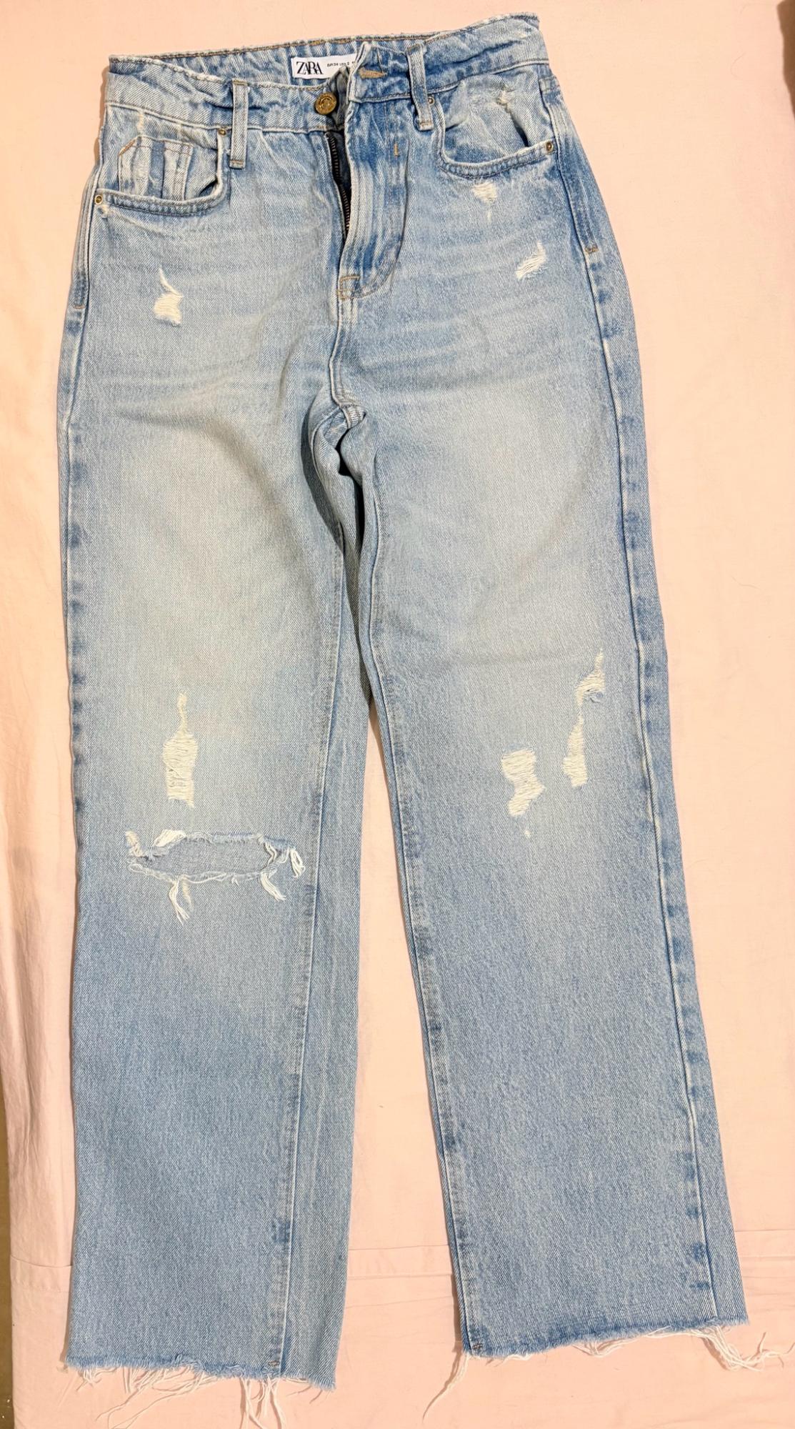 Light blue distressed jeans with a frayed hem on a pink background. A label with the text ZARA is visible on the waistband.