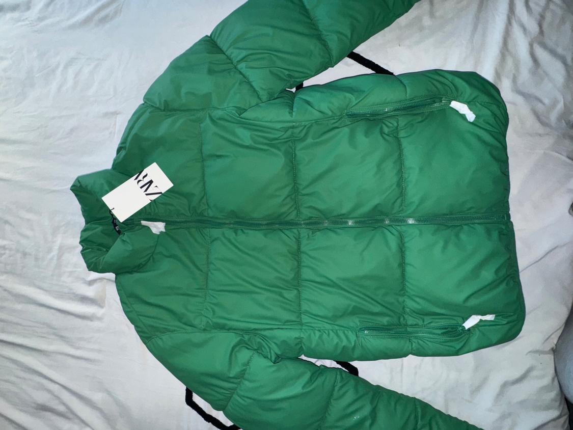 Green puffer jacket on a white bed, with a visible tag that says "ZARA".