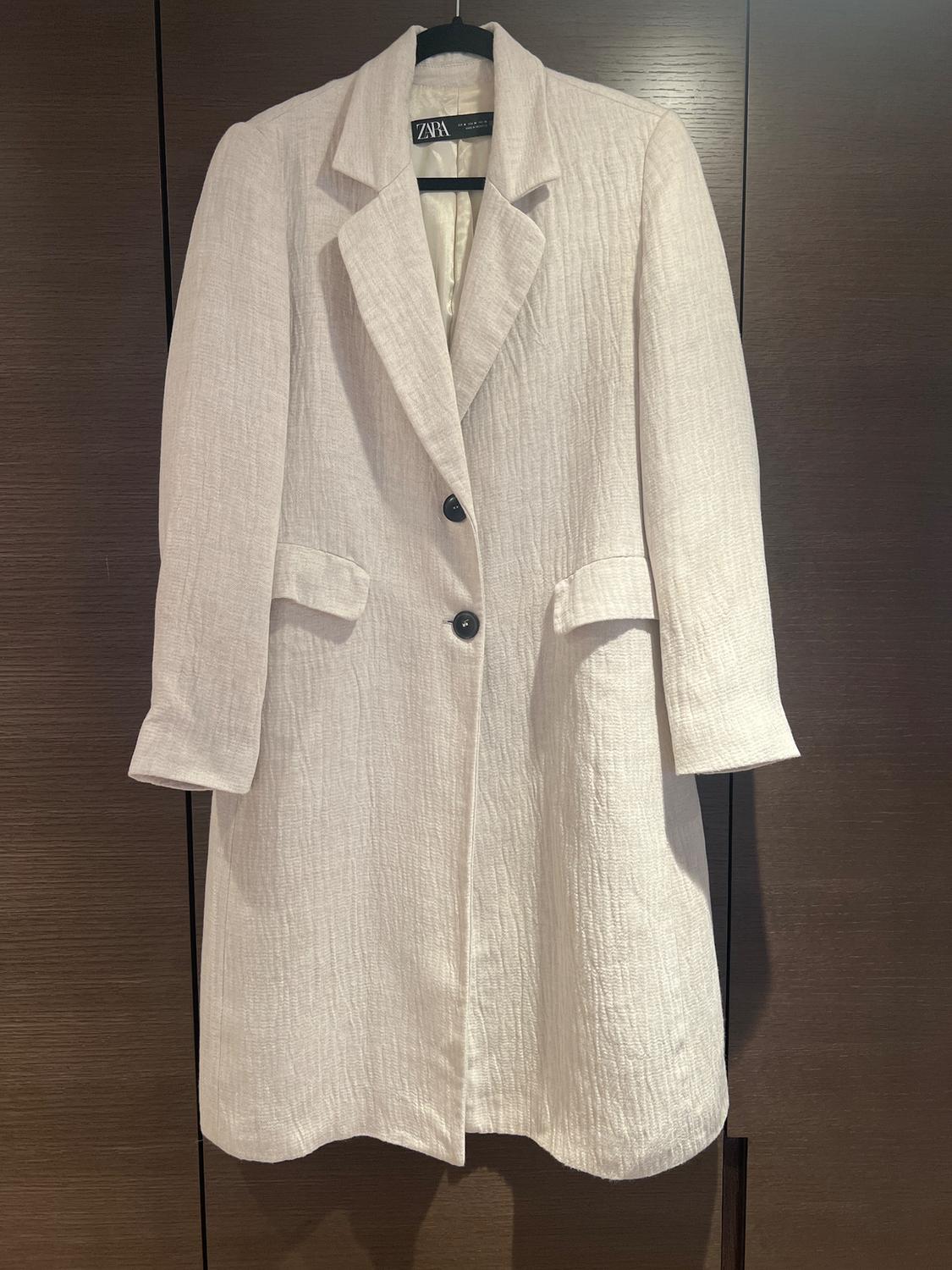 White textured fabric coat with two black buttons, hanging on a hanger in front of a dark wood background.