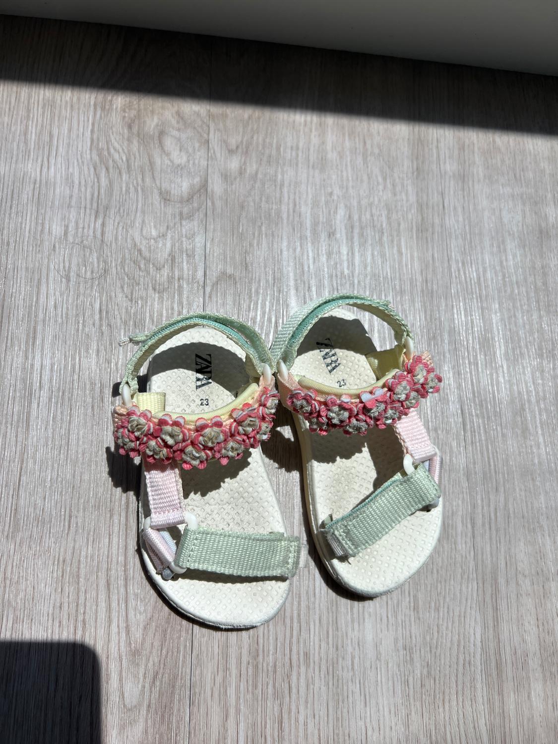 Children's sandals with embroidered flowers on the strap, pastel colours and a white sole, on a sunlit wooden floor.