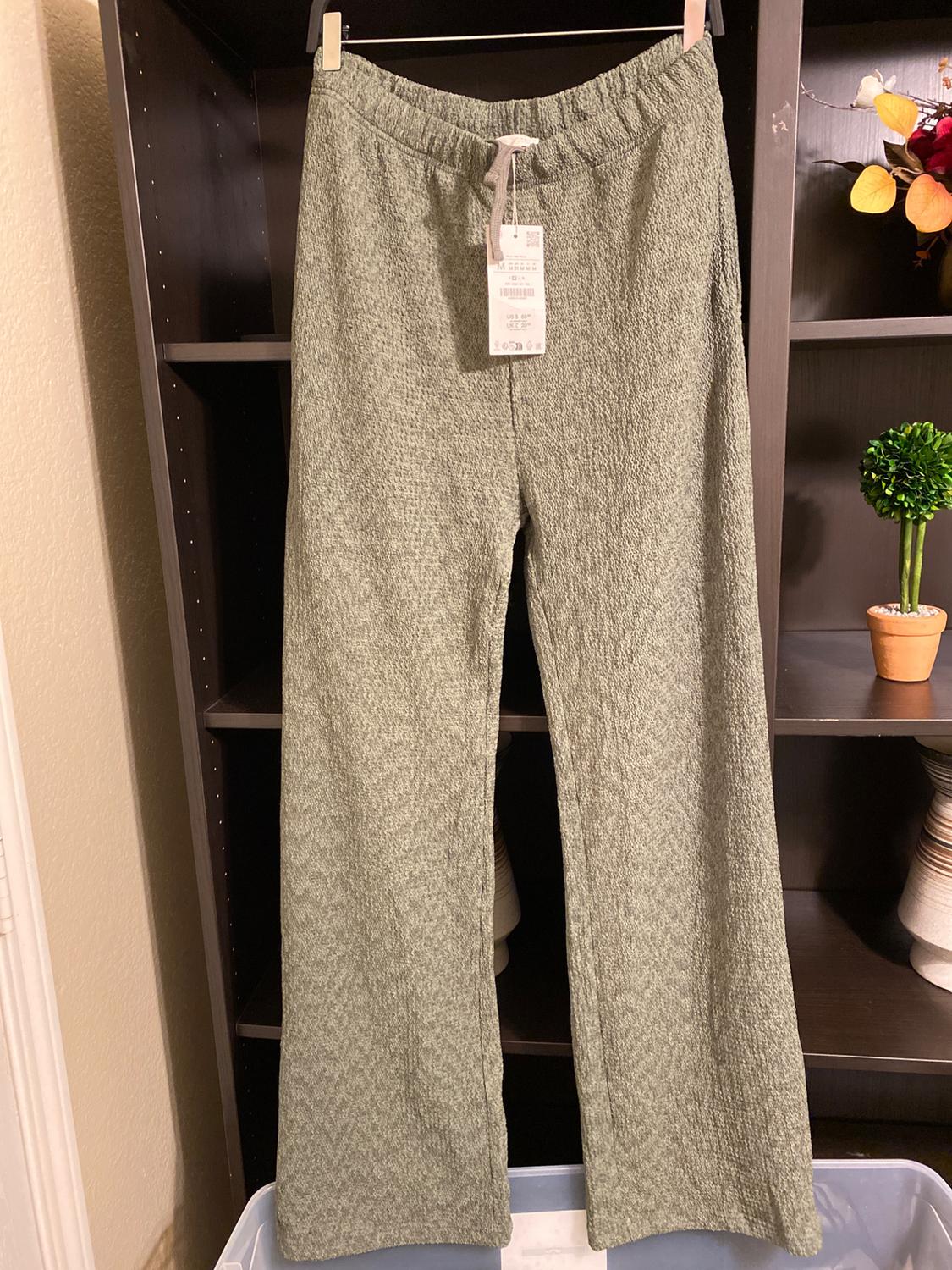 Green trousers hanging on a dark shelf, with a visible label. Background decorated with artificial plants and flowers.