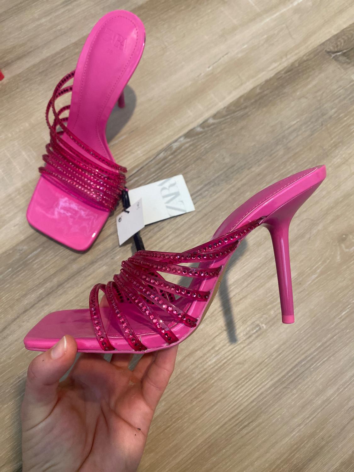 Pink high-heeled shoe with glittery straps, held by a hand over a light wooden floor. Label visible.