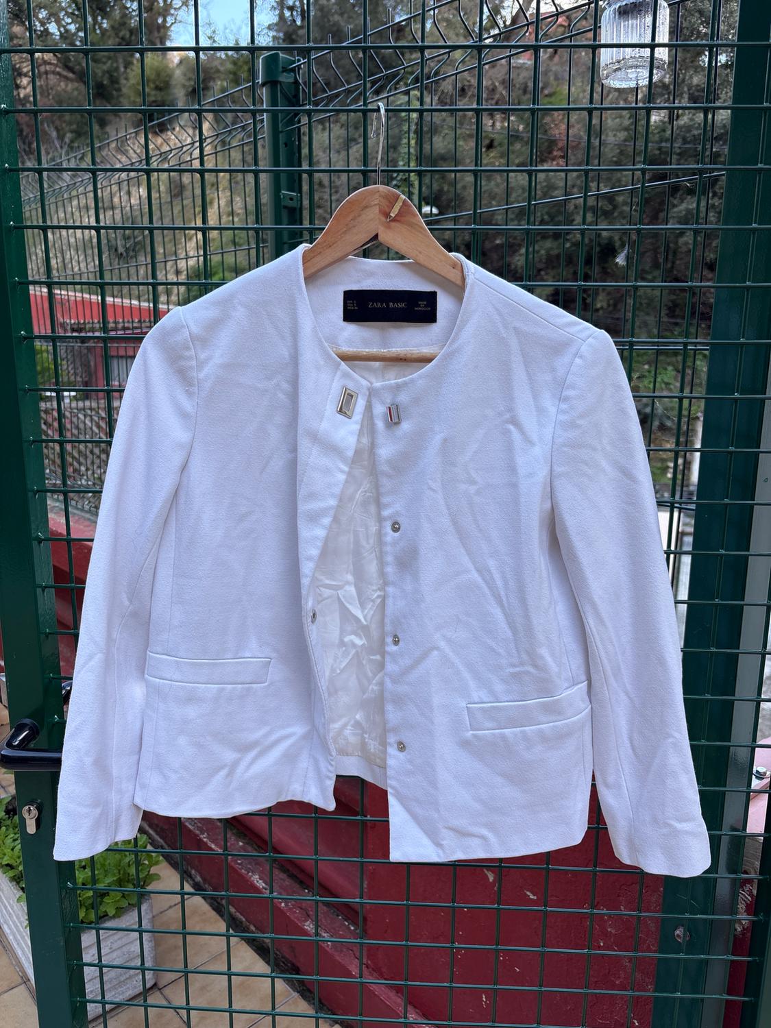 White women's jacket hanging on a wooden hanger against a green railing. Visible label: "ZARA BASIC". Garden background.