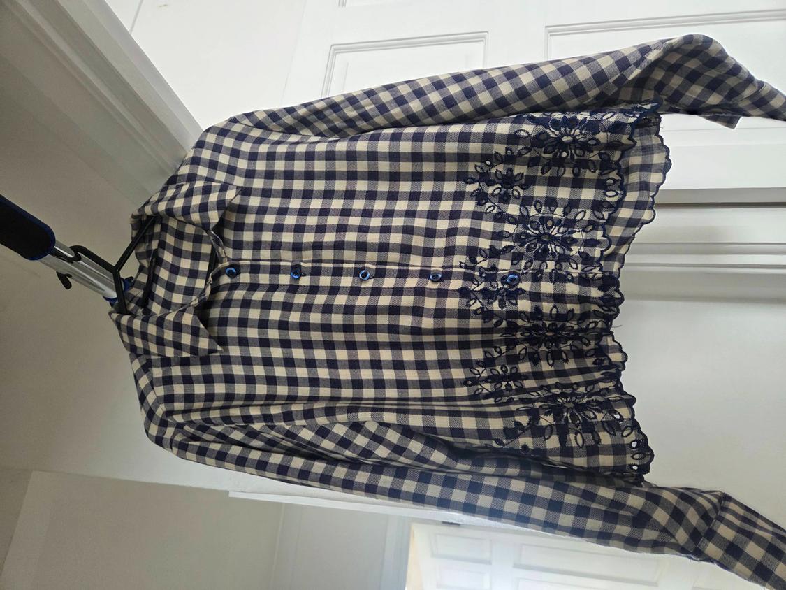 Blue and white checked shirt with floral embroidery on the hem, hanging on a hanger in front of a white door.