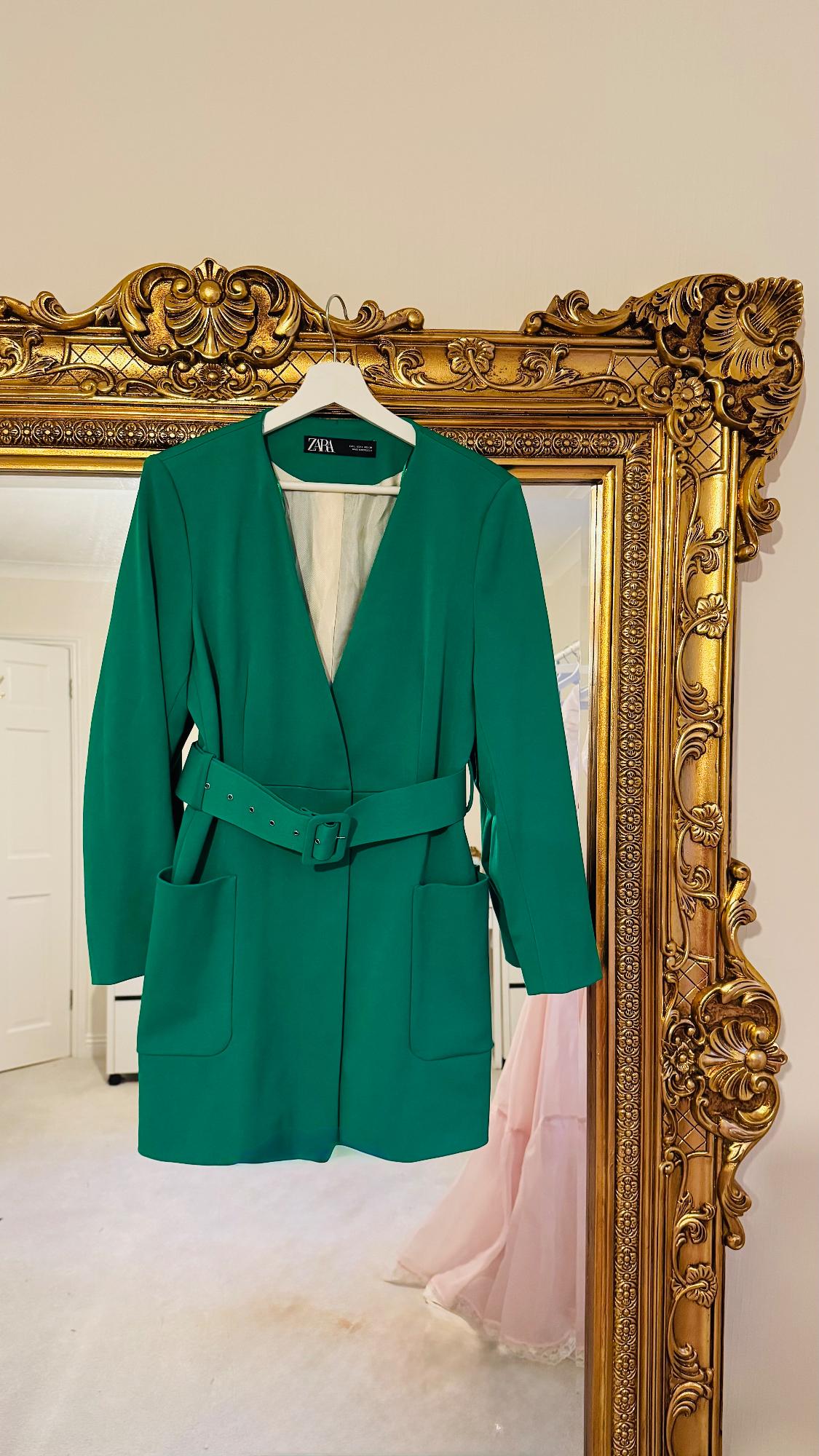 Green belted jacket hanging on an ornate gold mirror. Visible label: ZARA. Light-coloured room in the background.
