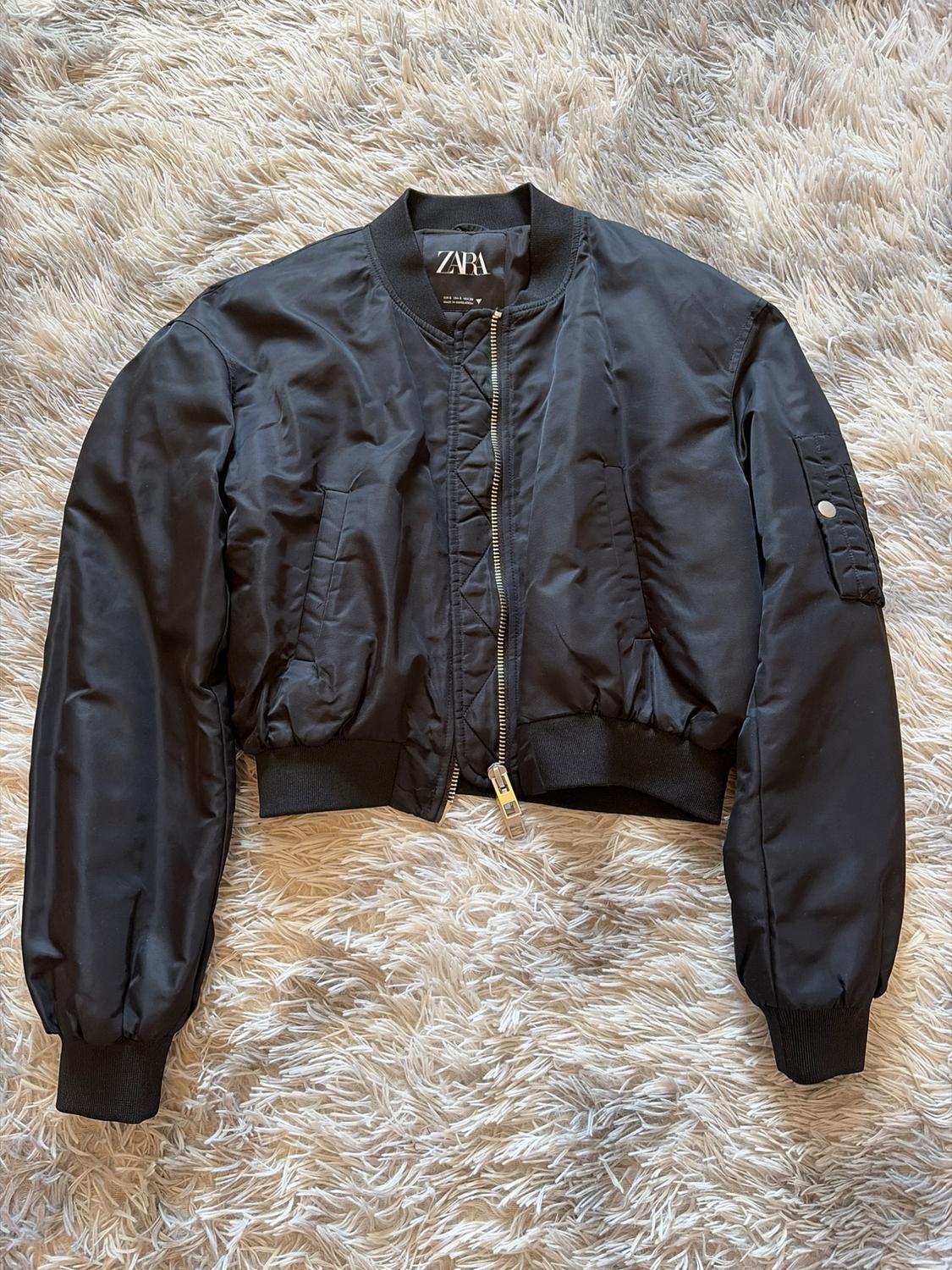 Μαύρο bomber jacket με φερμουάρ και ελαστικές μανσέτες, πάνω σε μπεζ χνουδωτό χαλί. Ορατή ετικέτα: ZARA.